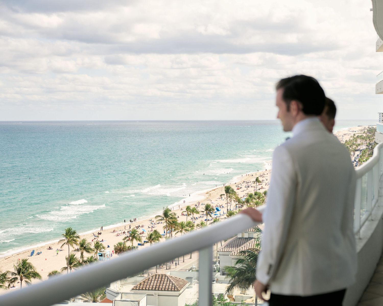 A person in white looks out over a tropical beach view from a high balcony.