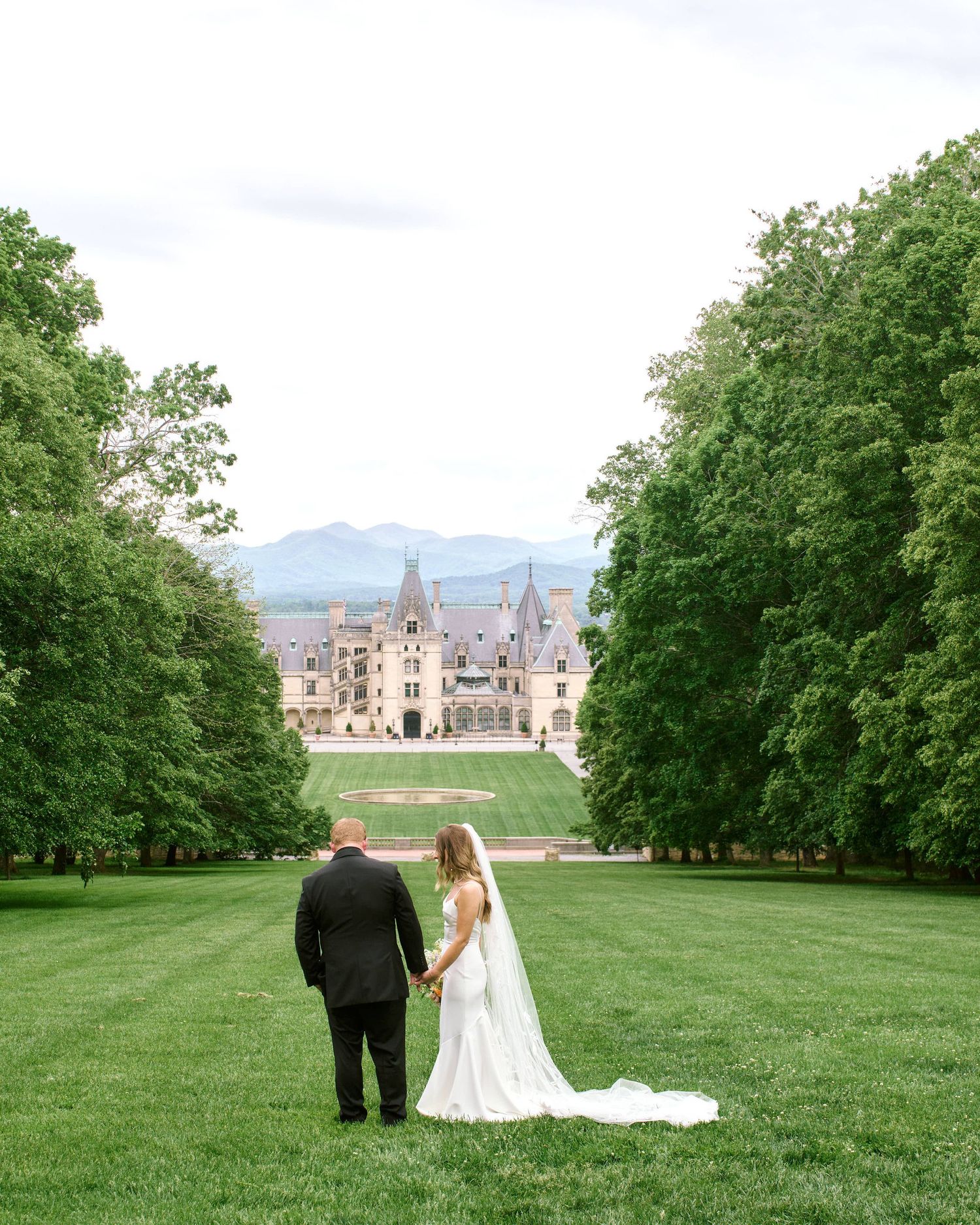A couple stands on a manicured lawn overlooking the grand Biltmore Estate mansion with mountains in the distance.