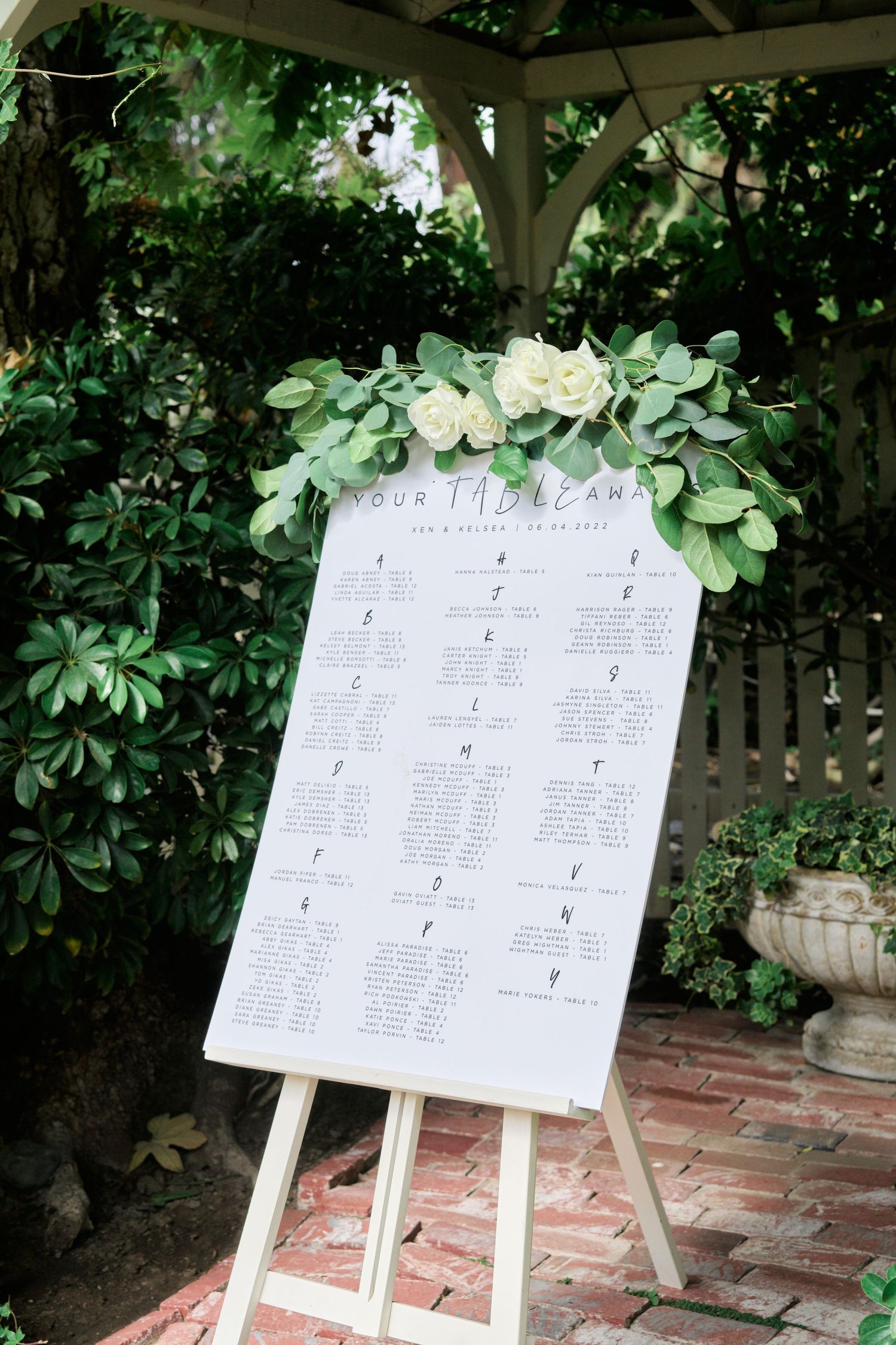 Wedding seating chart displayed on an easel decorated with white flowers and greenery in a garden setting.
