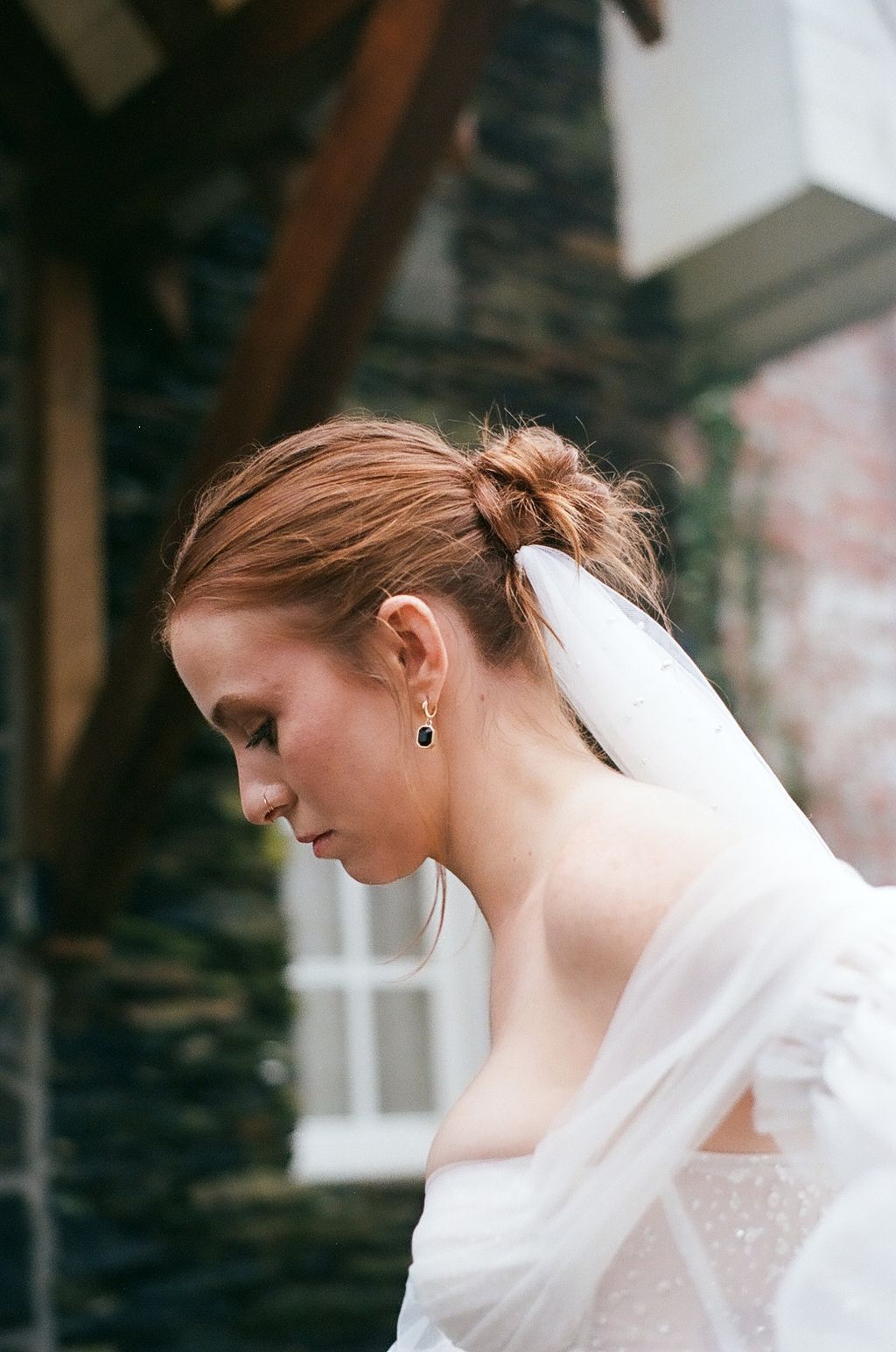Side profile of bride wearing delicate veil and earrings against stone wall background.