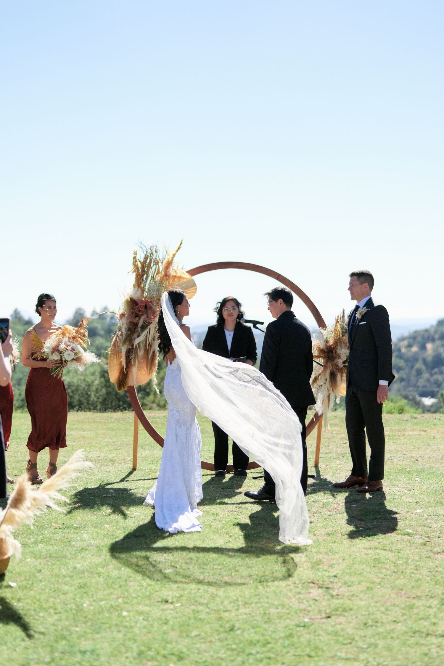 Wedding ceremony under a circular arch with dried pampas grass decorations on a hilltop.