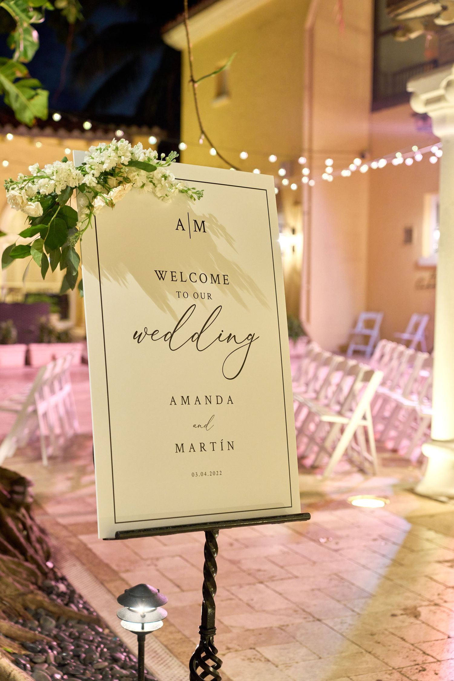 Elegant wedding welcome sign with floral decor and string lights in outdoor venue setting.