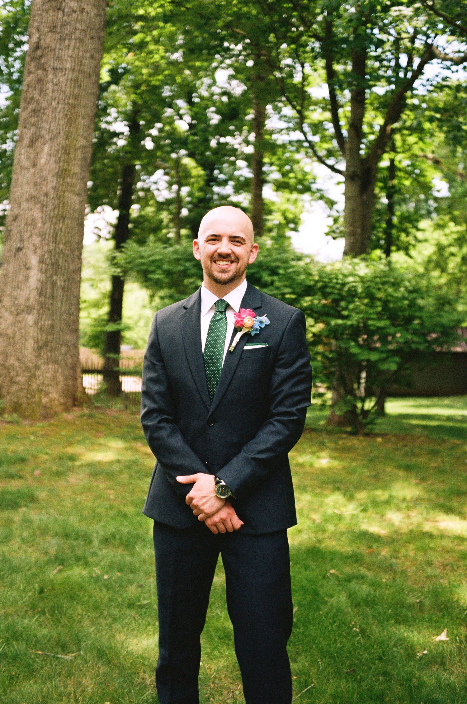 Smiling bald groom in dark suit with green tie and boutonniere standing outdoors on green lawn.