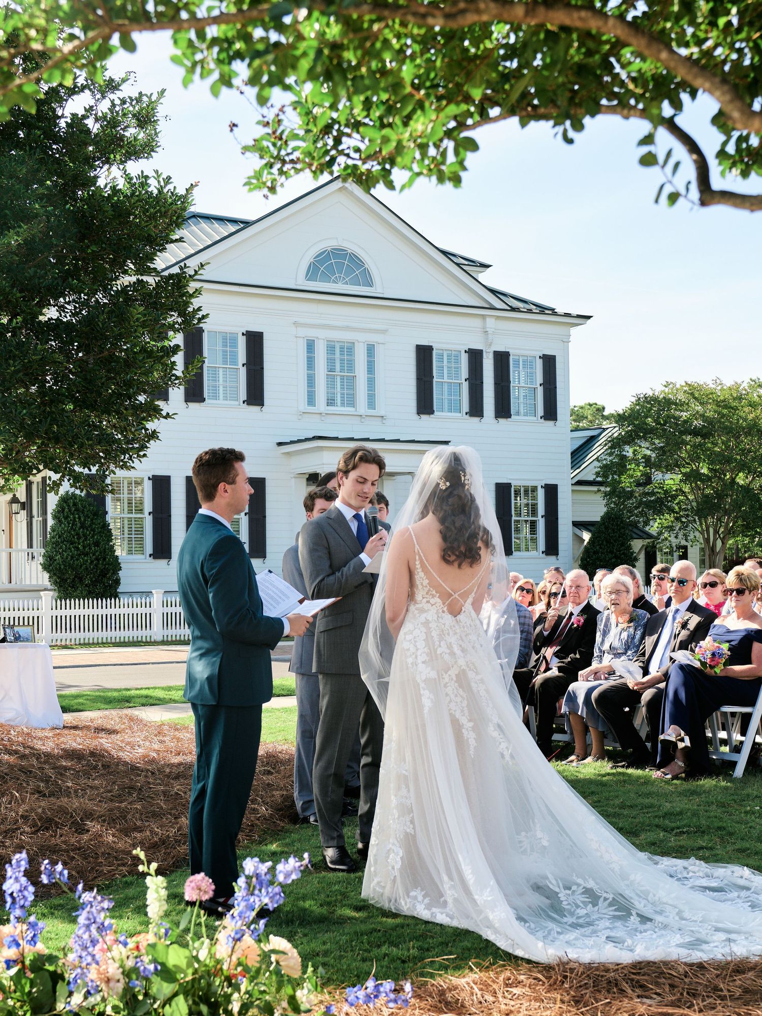 Outdoor wedding ceremony on lawn before white colonial mansion, bride in lace gown with veil, groom in gray suit, guests seated.