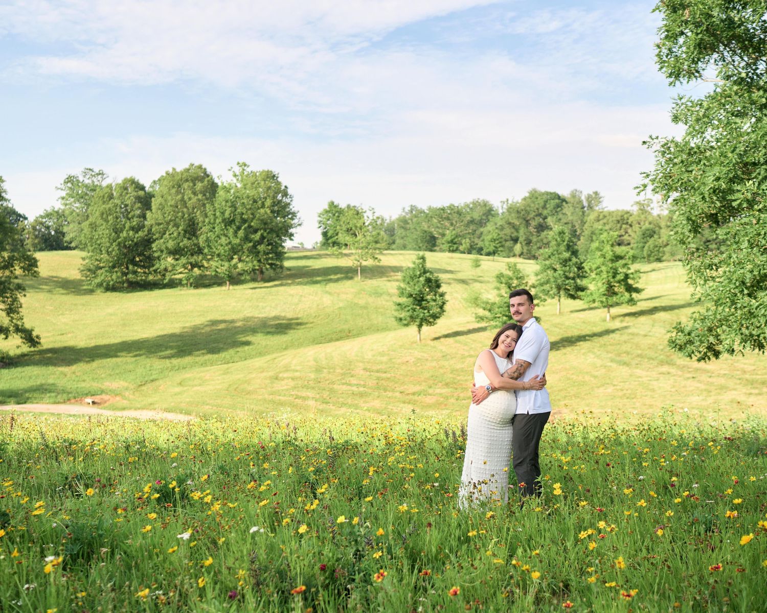 A couple embraces in a lush green meadow with wildflowers, trees, and rolling hills on a sunny day.