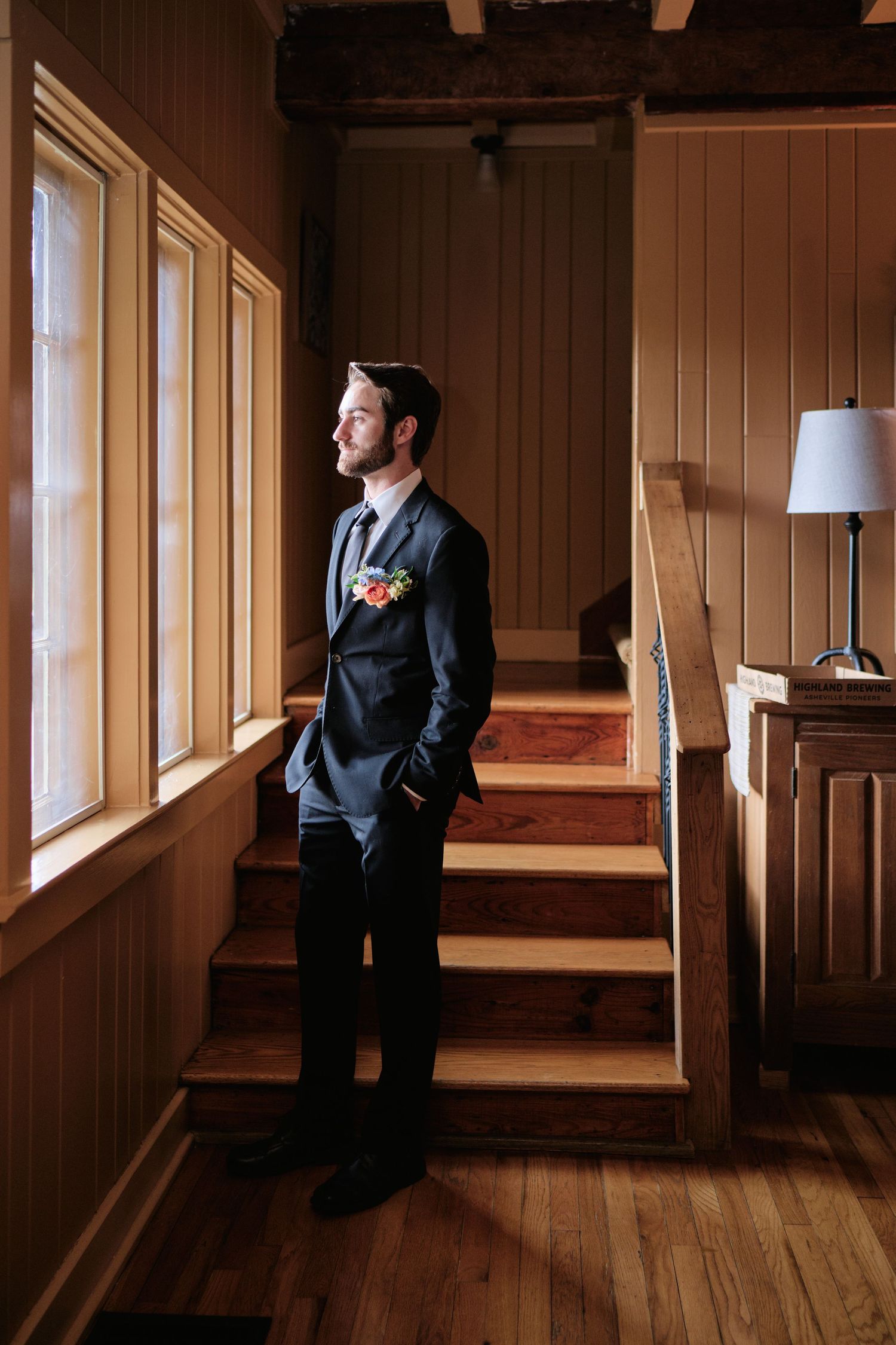 Groom in dark suit with floral boutonniere stands on wooden stairs by windows in rustic wood-paneled venue.