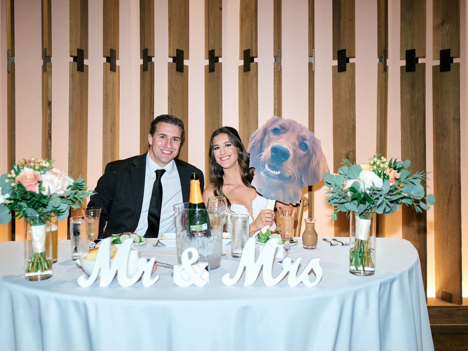 Bride and groom pose at sweetheart table with Mr & Mrs sign, floral arrangements, and their dog.