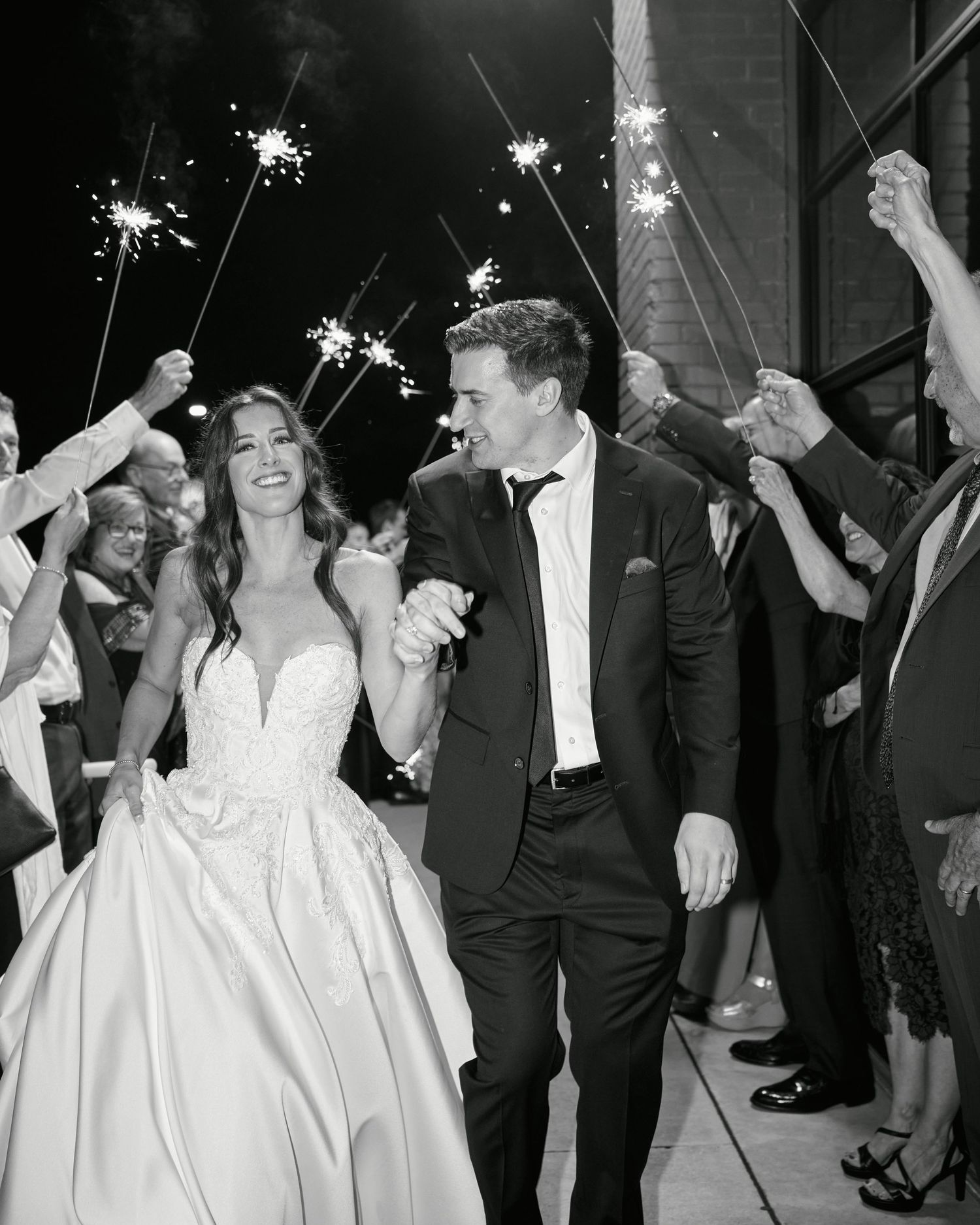 Black and white photo of a joyful bride and groom walking through a sparkler send-off at their wedding reception.
