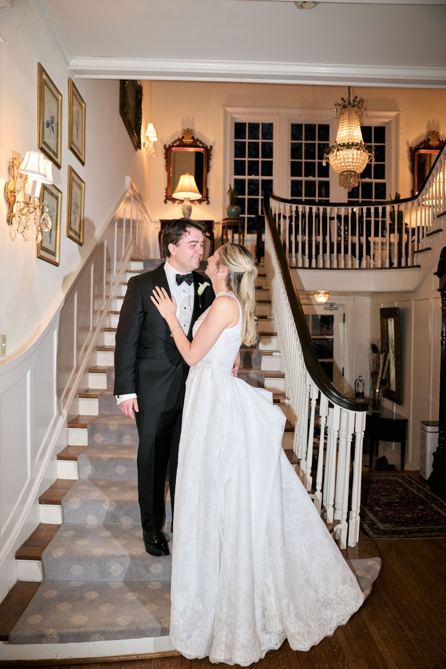 Bride and groom share a romantic moment on elegant staircase in a grand venue, bride in lace gown, groom in black tuxedo.