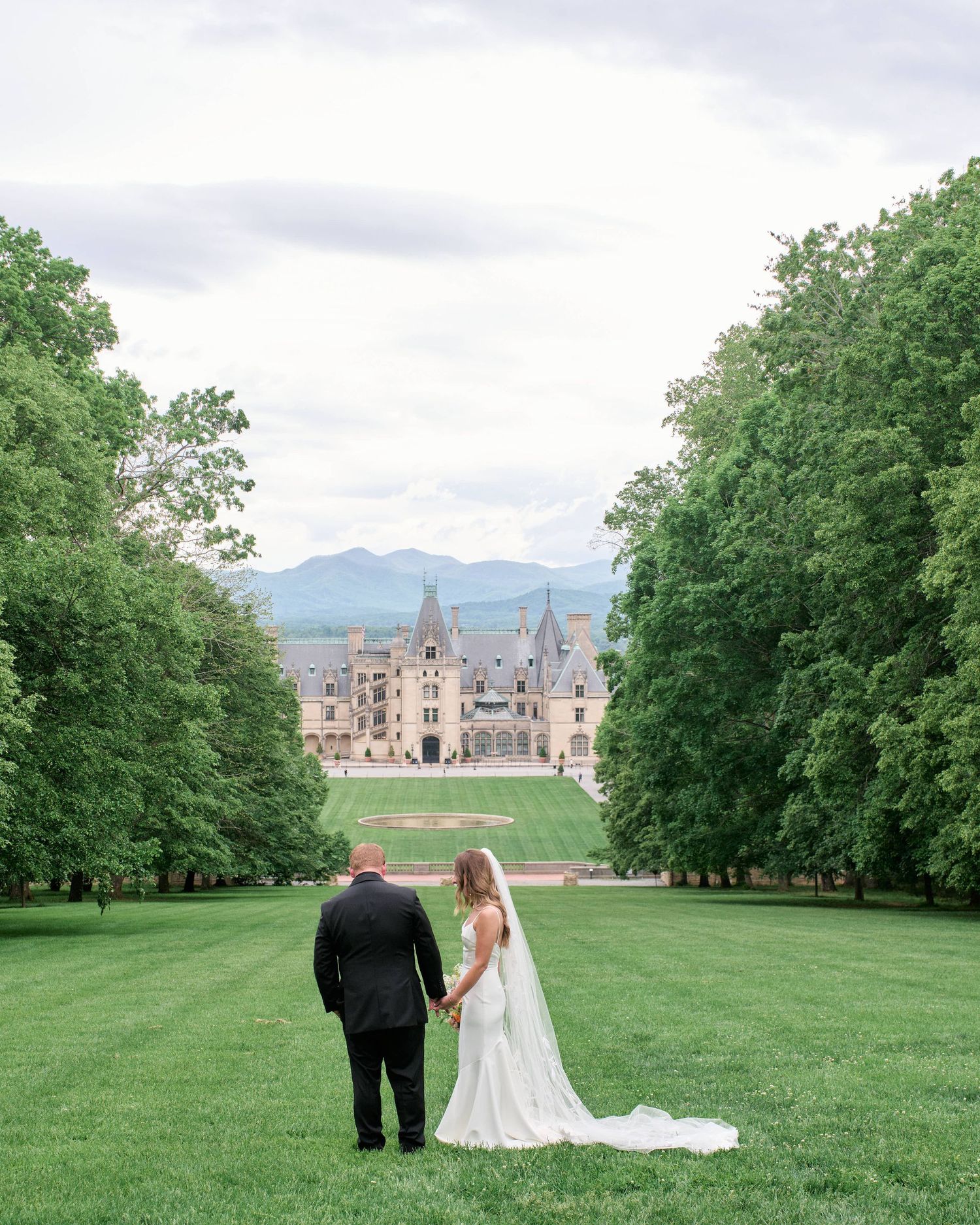 A couple walks hand in hand across a lush green lawn towards a majestic castle-like building in the distance.