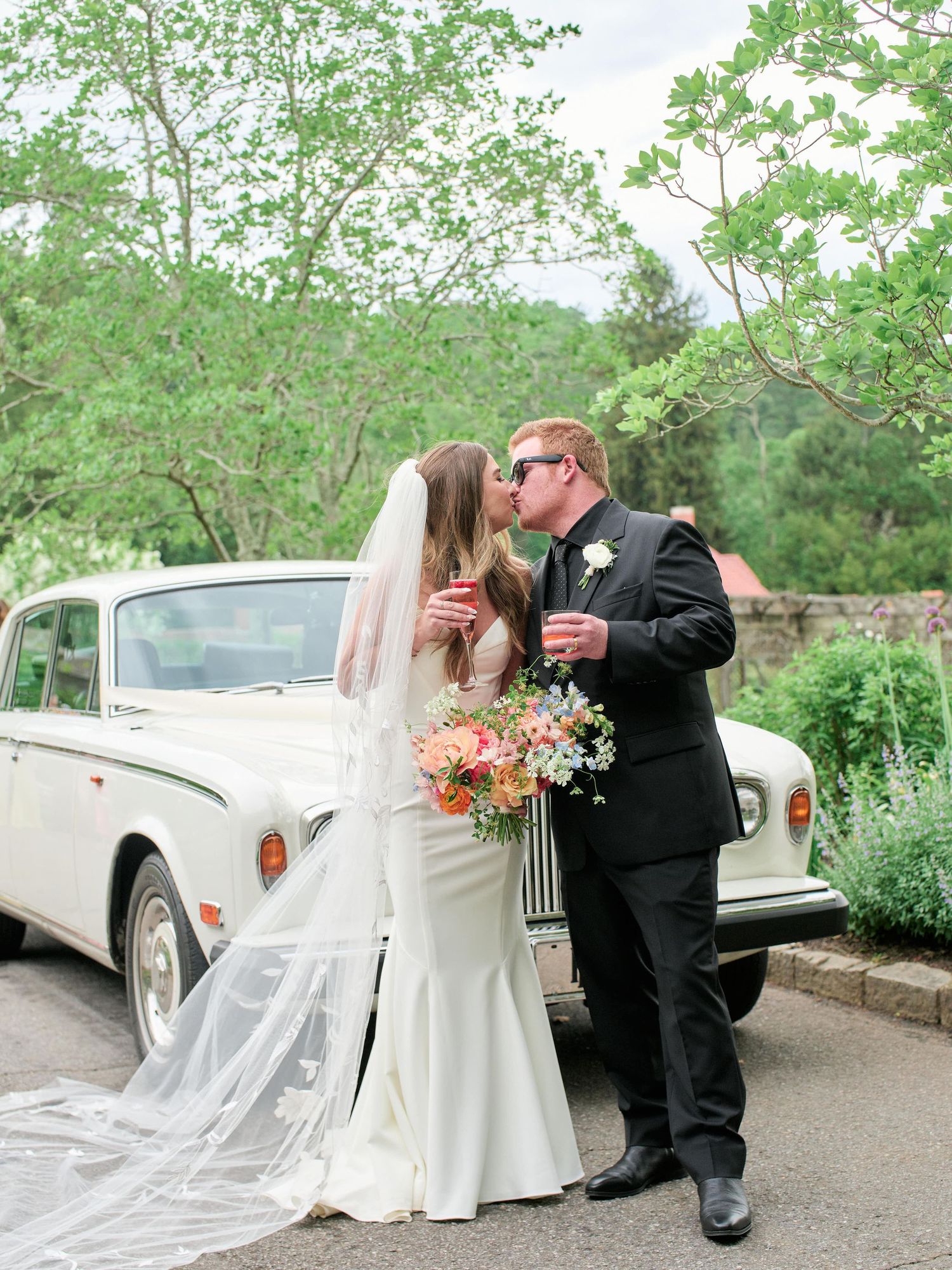 A romantic wedding couple shares an intimate moment next to a vintage white car in a lush garden setting.