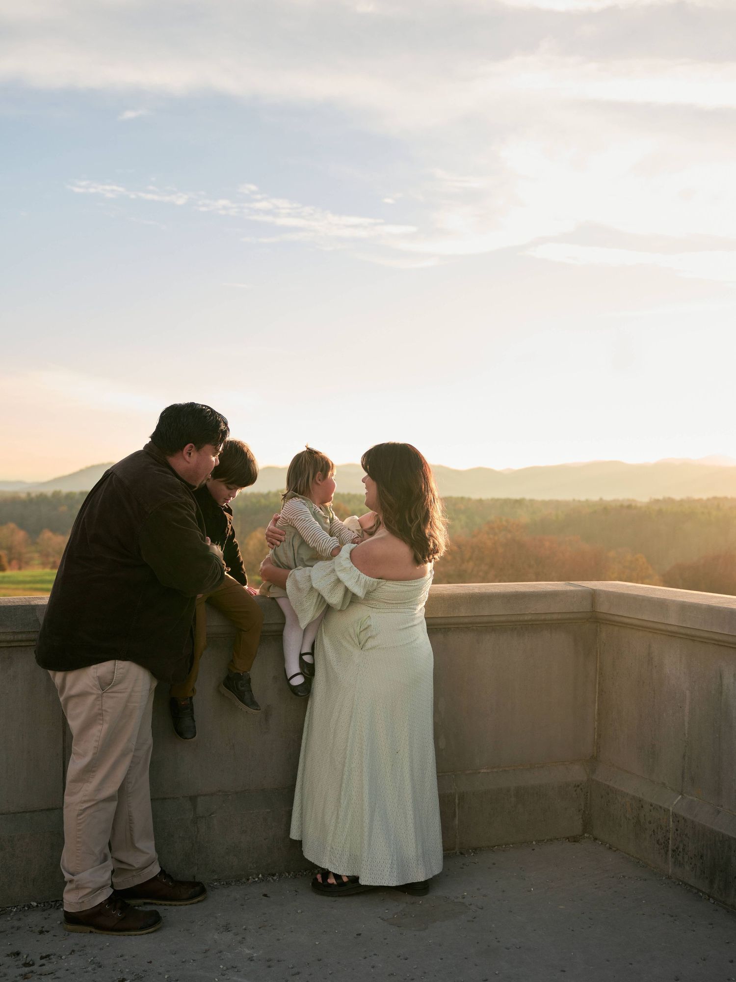 Family of three sharing a tender moment on a stone terrace at golden hour with rolling mountain views.