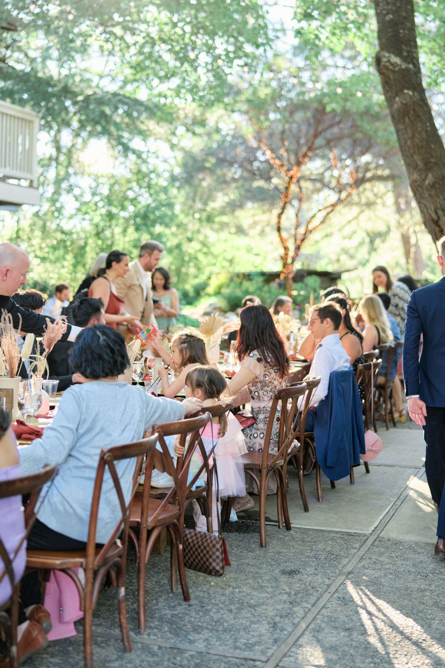 Outdoor wedding reception with guests seated at long tables under trees.