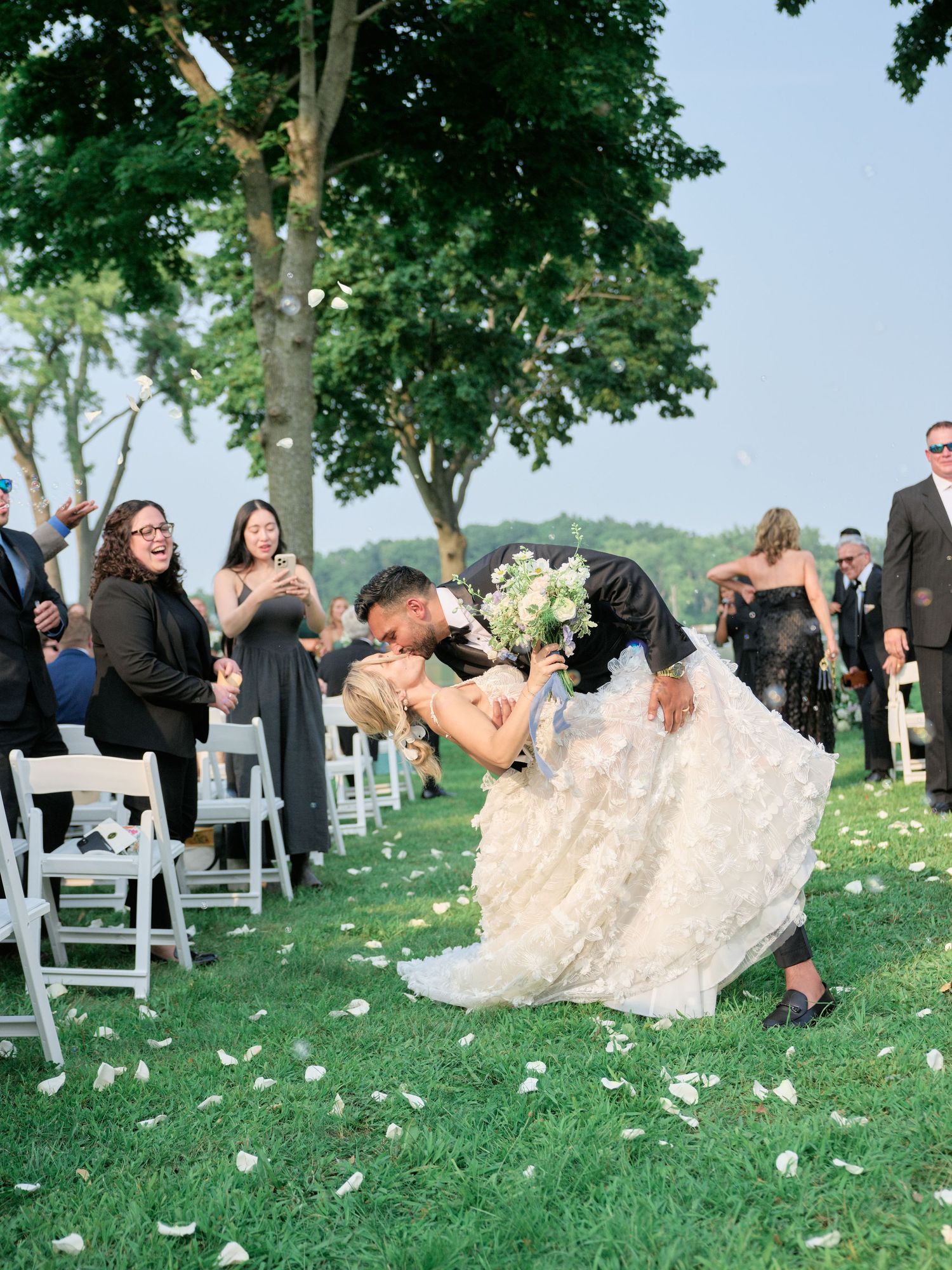 Wedding guests celebrate joyfully during outdoor garden ceremony on green lawn with white chairs and floral petals.
