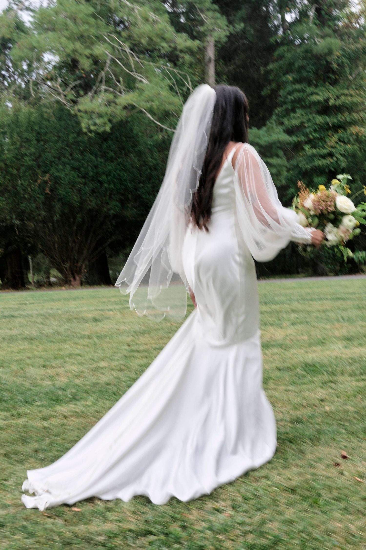 Bride in flowing white dress and veil twirls on grass at outdoor wedding.