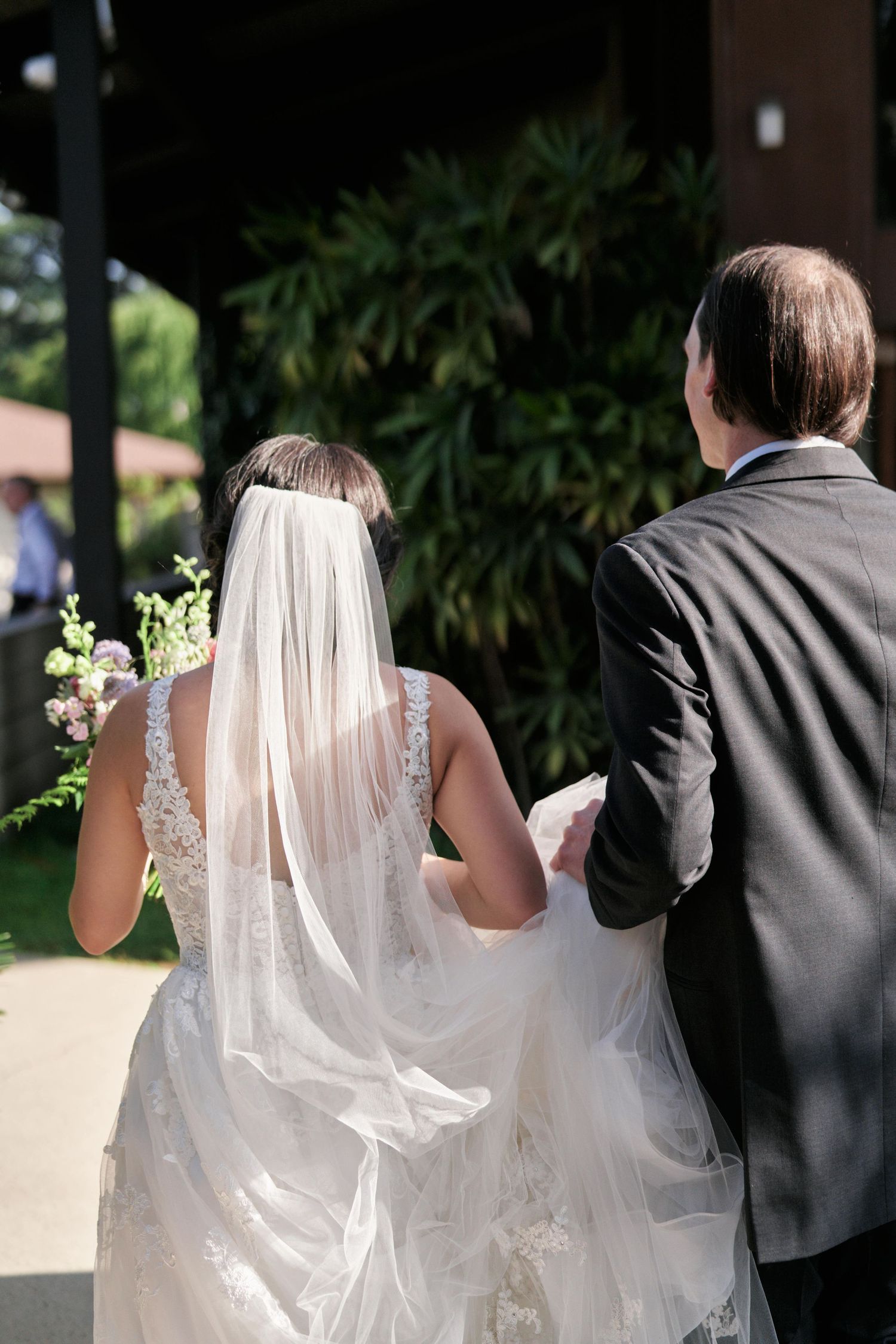 A bride in lace dress with flowing veil walks outdoors on her wedding day.