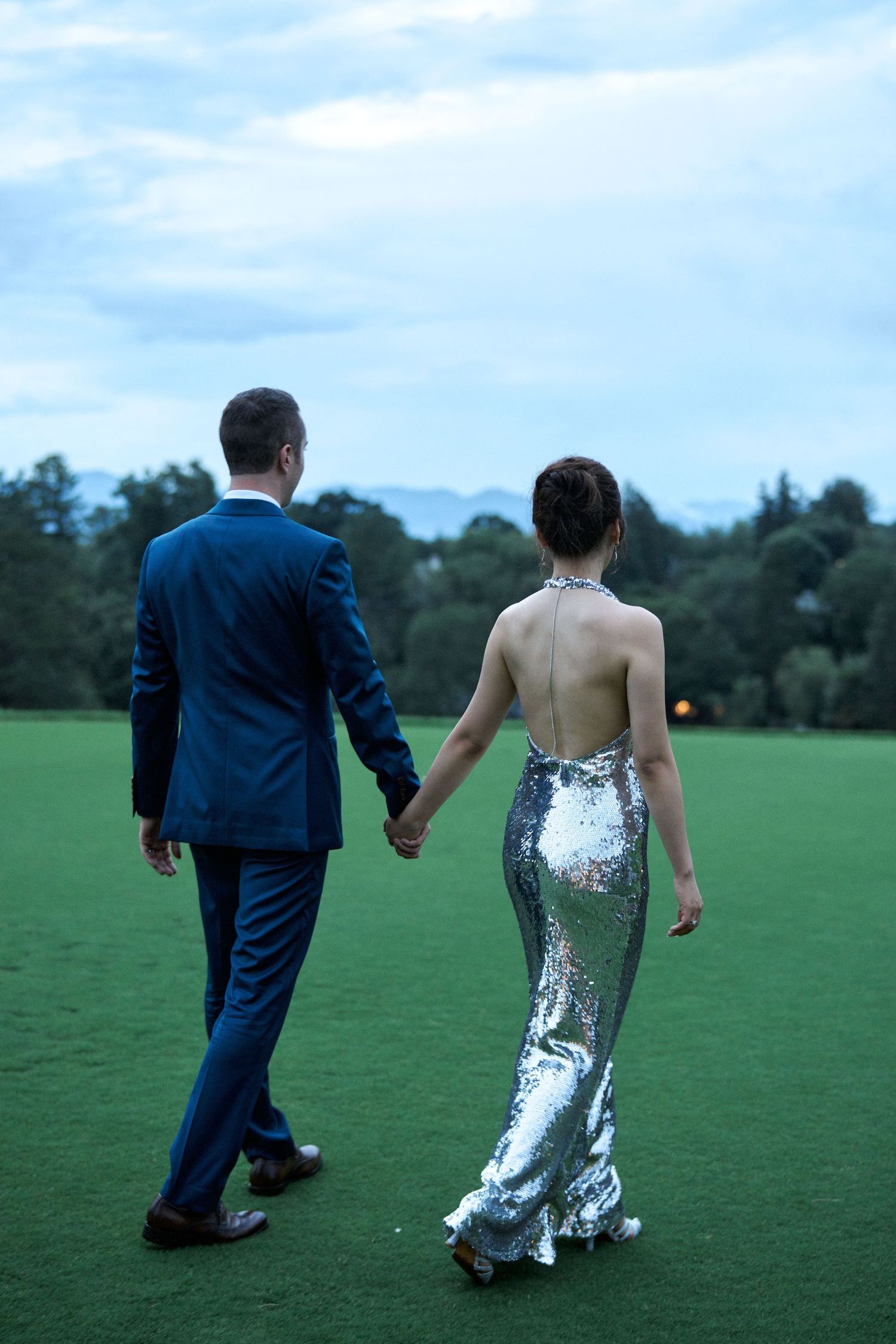 Couple walks hand in hand across a green lawn, bride wearing a sparkly silver dress.
