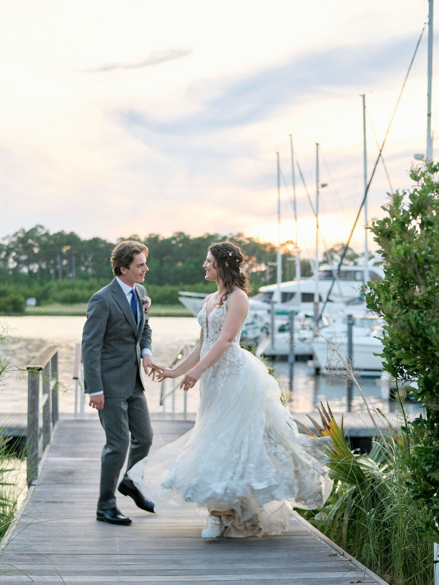 Newlywed couple stands together on a marina dock at sunset with boats and water in the background.