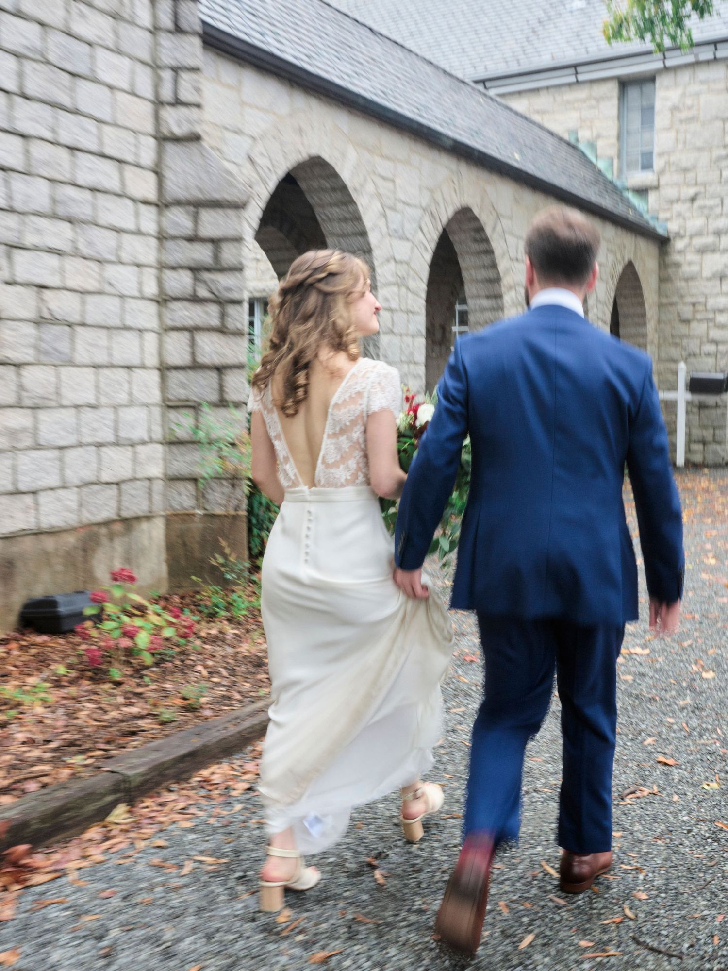 A couple in wedding attire walks hand in hand past stone arches of a historic church building.