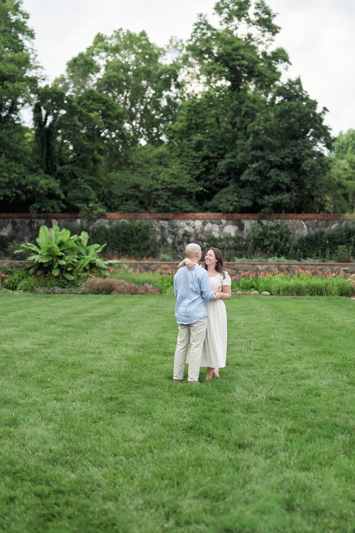 A couple embraces on a manicured lawn with stone walls and gardens in the background.