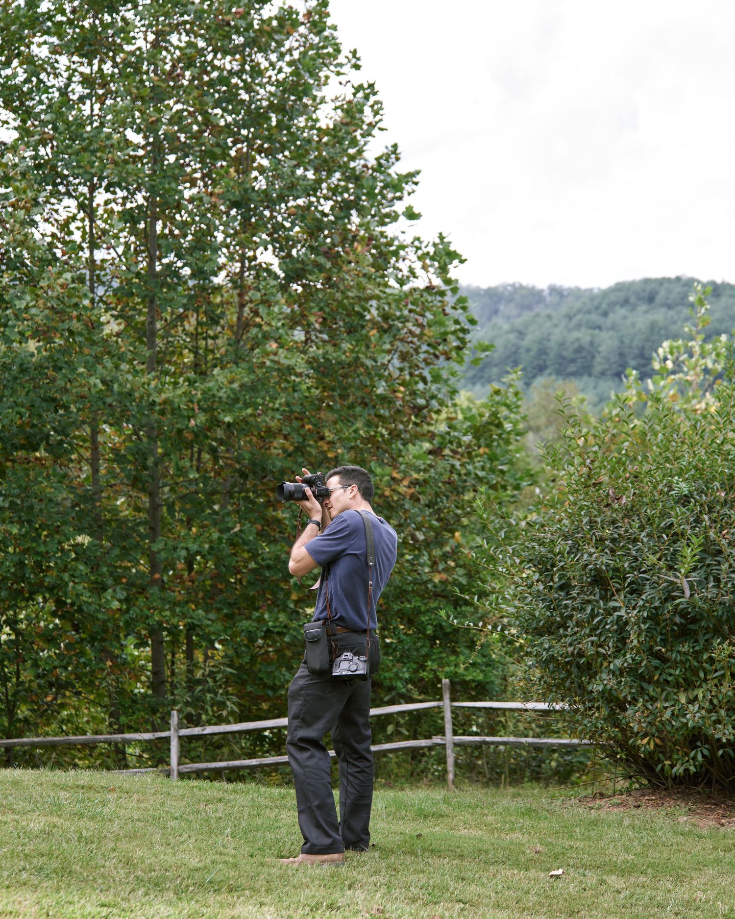 Photographer in dark clothing captures autumn scenery in an apple orchard with rolling hills in the background.