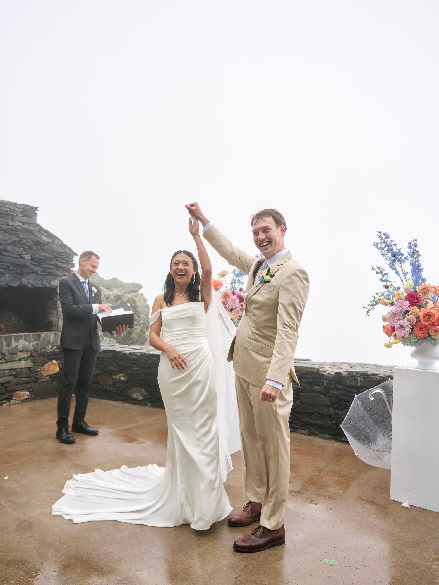 Joyful newlyweds celebrate at outdoor wedding ceremony with colorful floral arrangements on a misty coastal terrace.