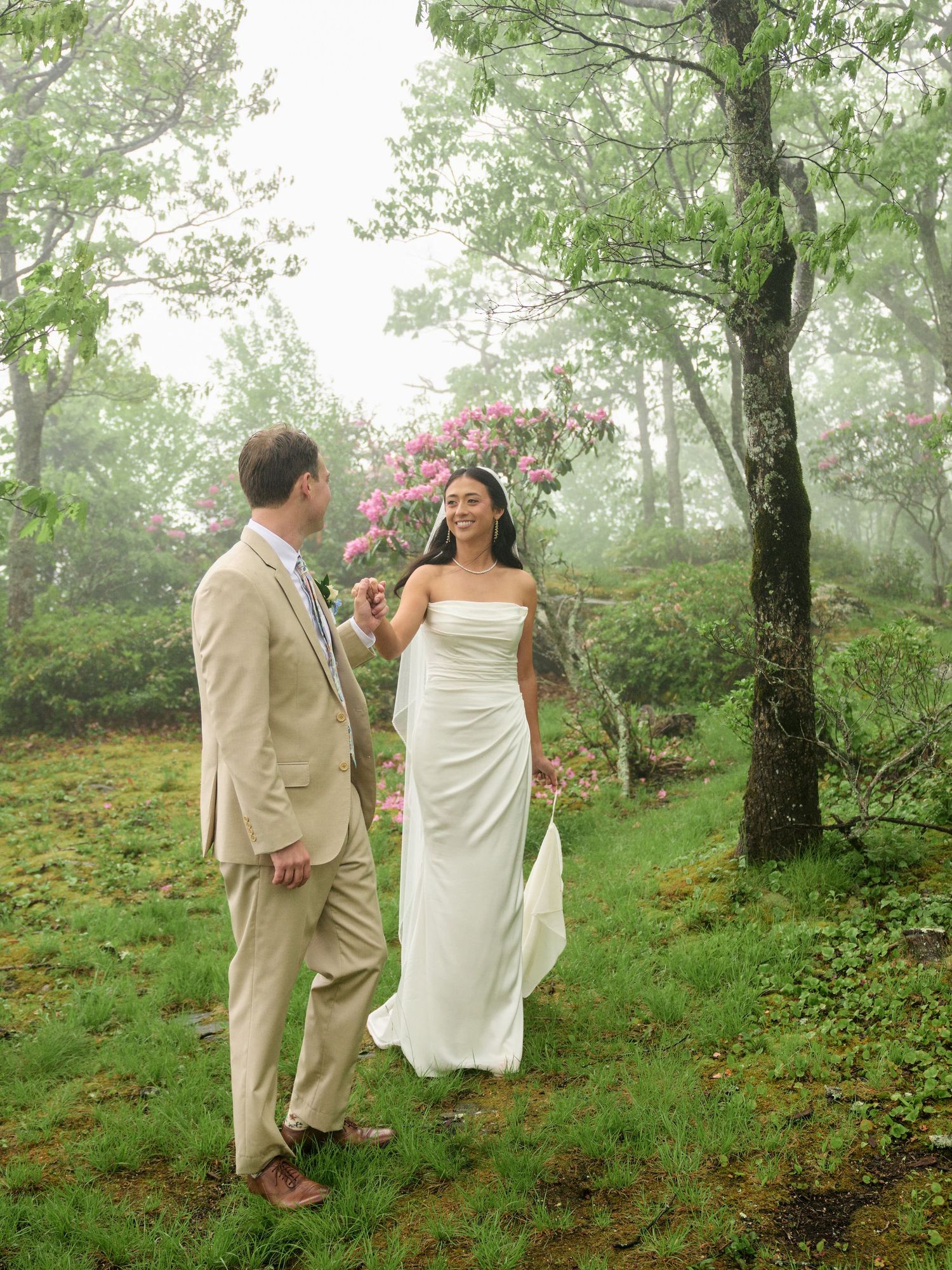 A bride in a white gown and groom in a tan suit share a joyful moment in a misty forest with pink blooms.