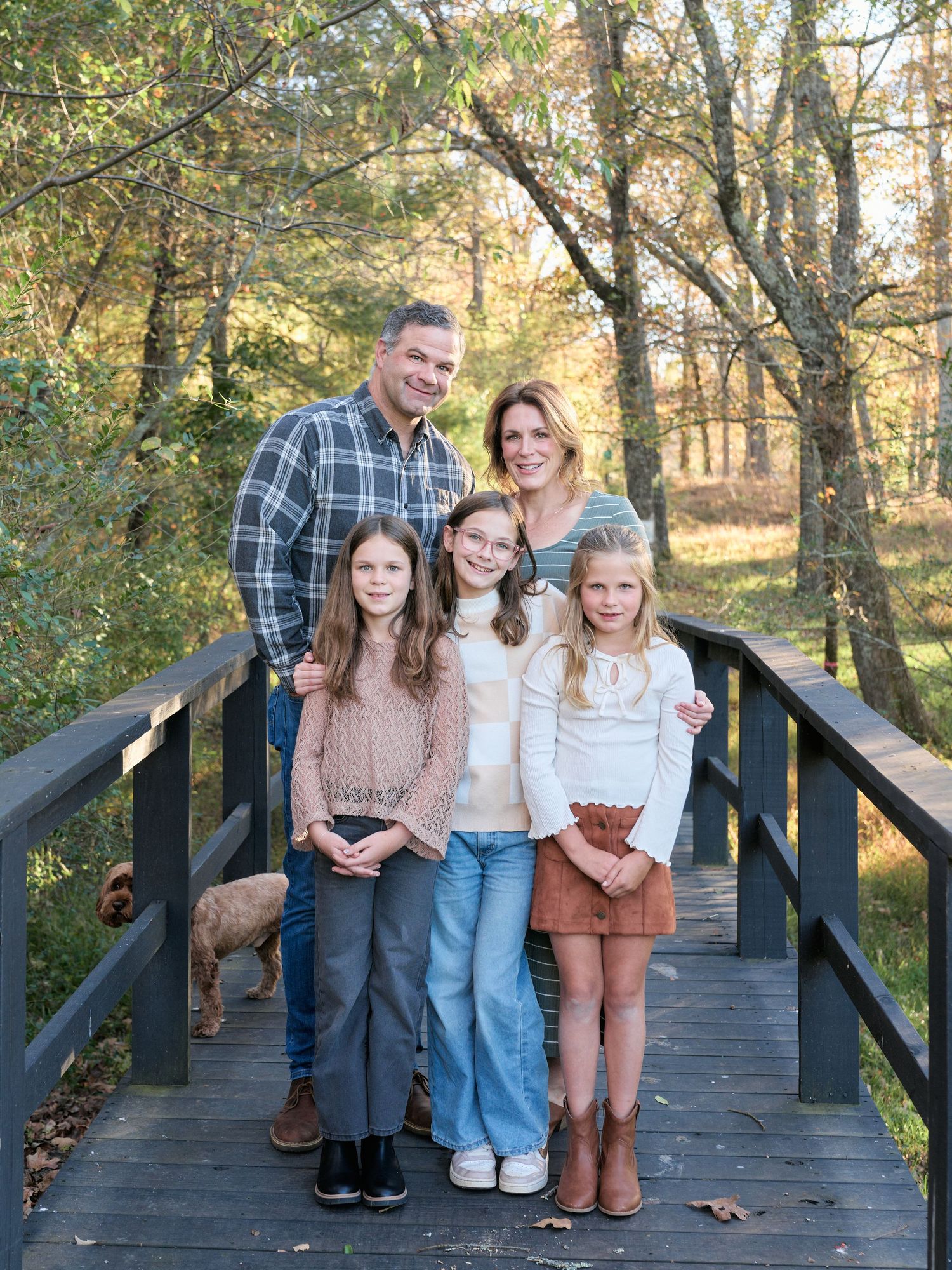 A family poses together on a wooden bridge in a wooded area during autumn.