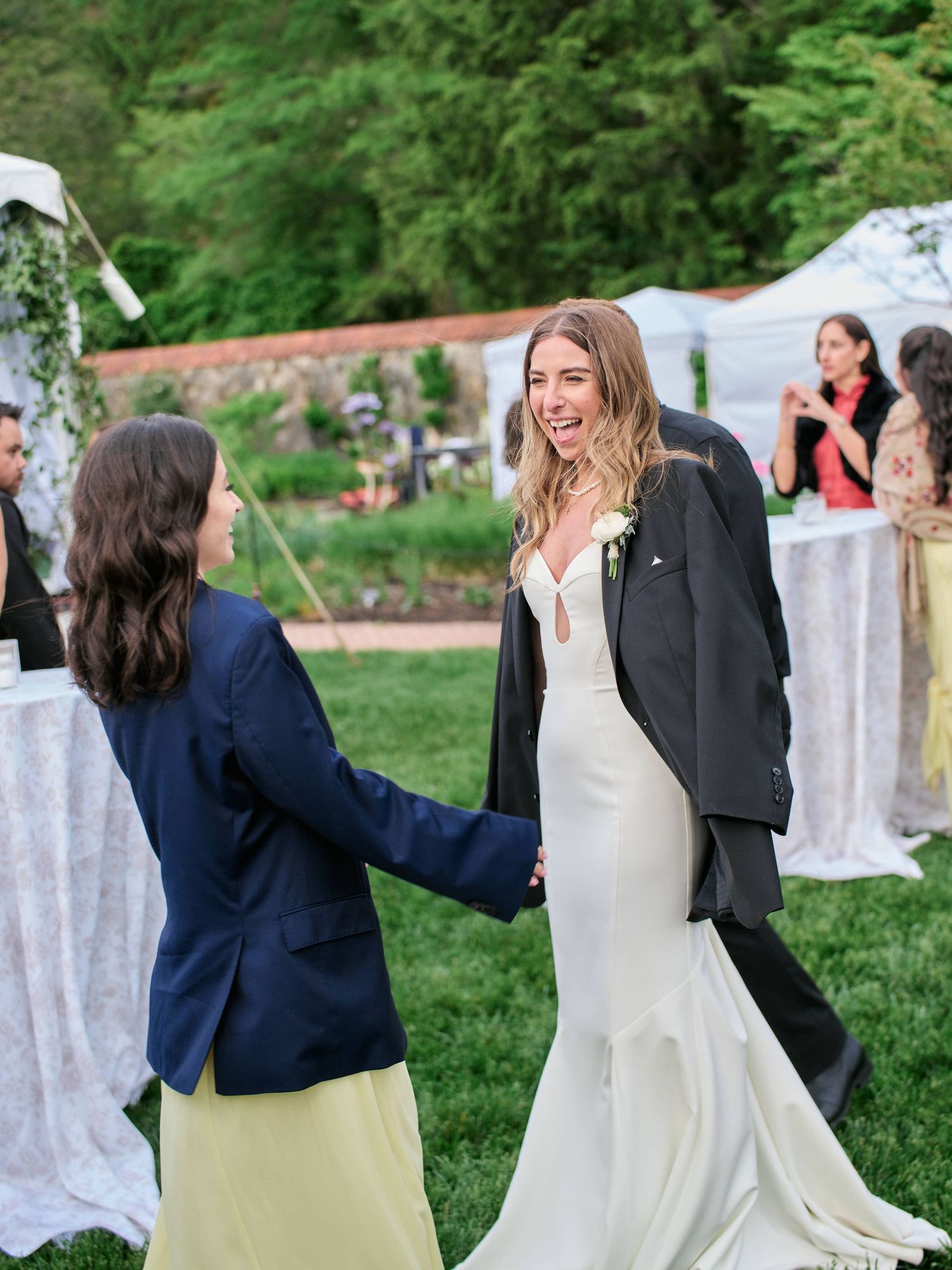 A bride in a white gown and someone in a navy suit dance together joyfully in an outdoor garden setting.