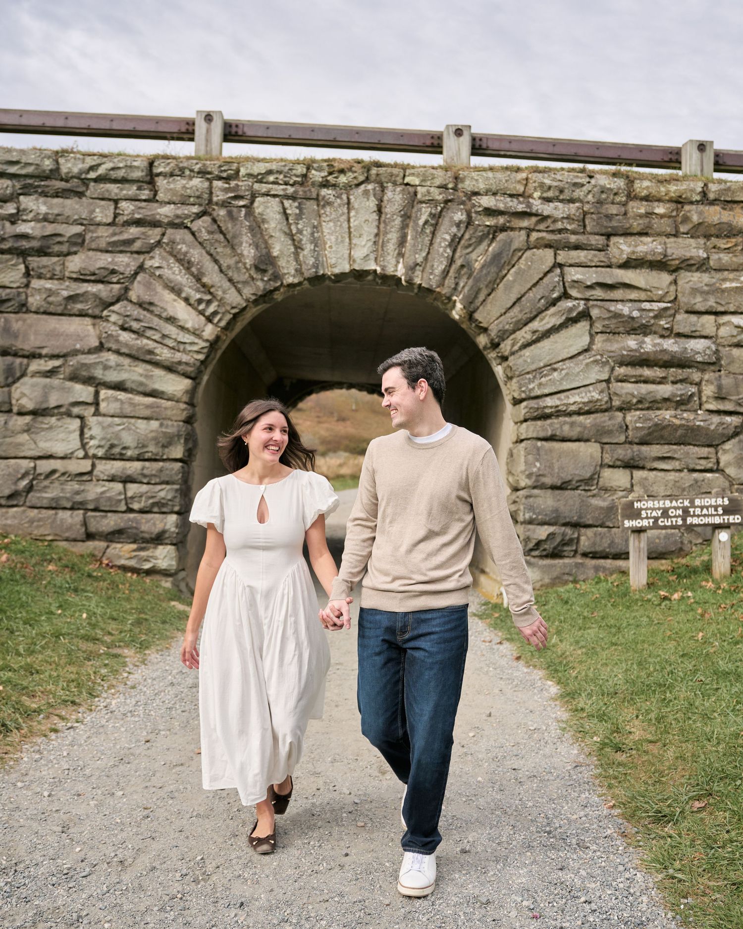 A couple walks hand in hand through a stone archway tunnel along a scenic path on a bright day.