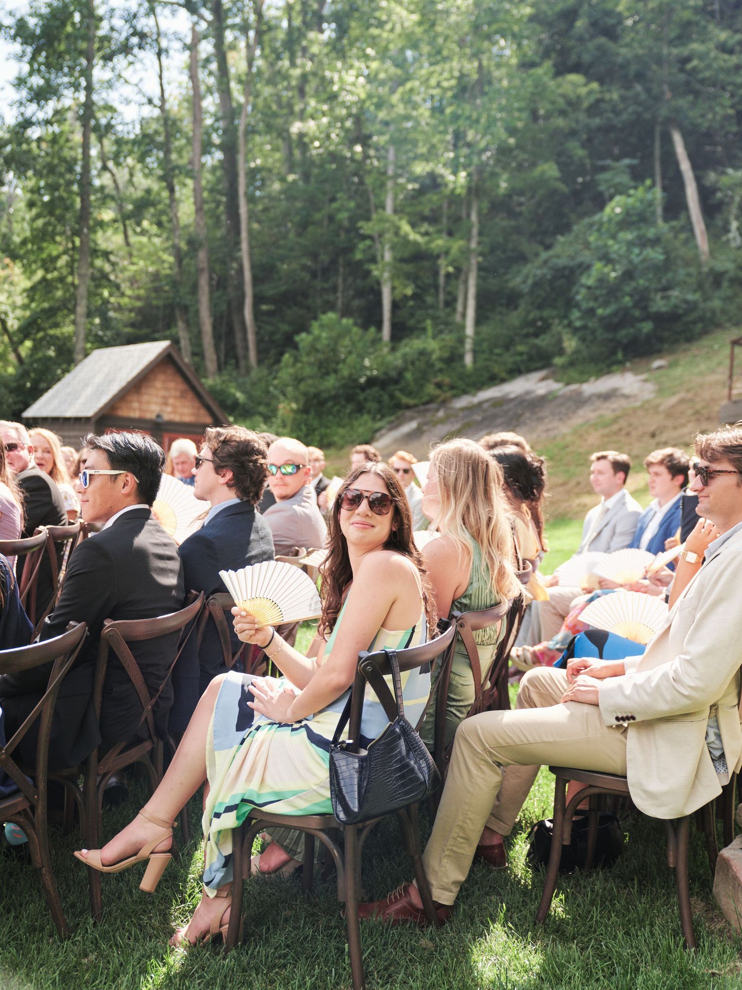 Guests seated outdoors at a sunny wedding ceremony, surrounded by lush green trees and a rustic barn backdrop.