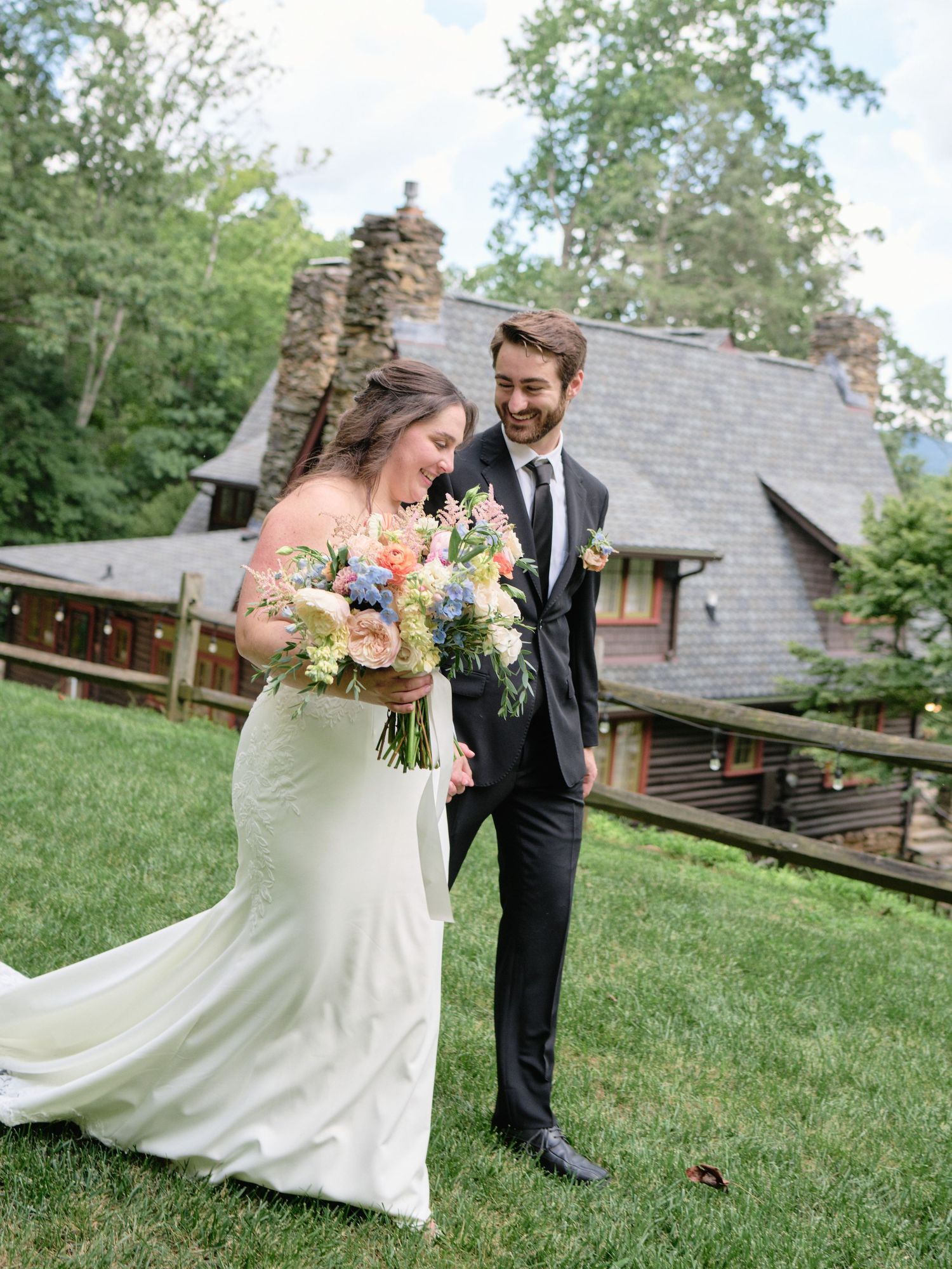 A smiling bride in a white gown holding a colorful bouquet stands with a groom in a black suit outdoors.