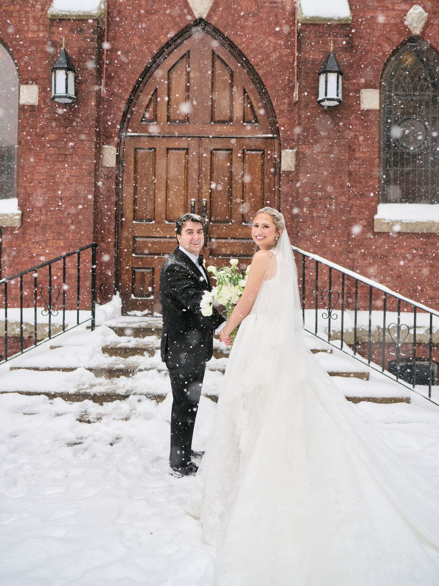 Bride and groom posing in snowfall on church steps in winter wedding photo with brick Gothic architecture backdrop.