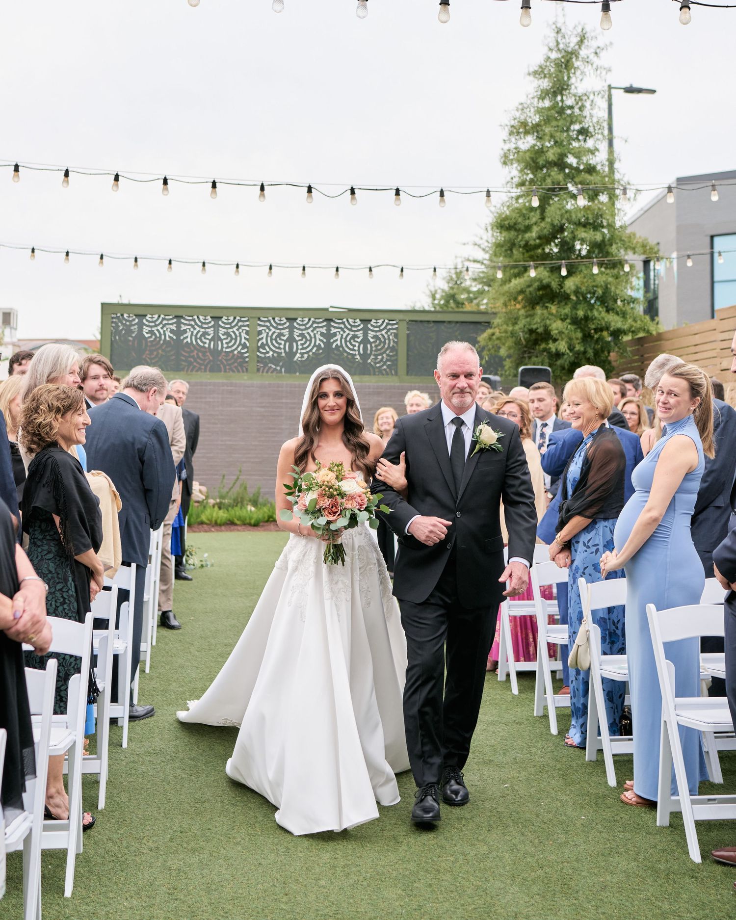 Bride walks down outdoor wedding aisle with escort, white chairs, string lights, and floral bouquet at rooftop venue.