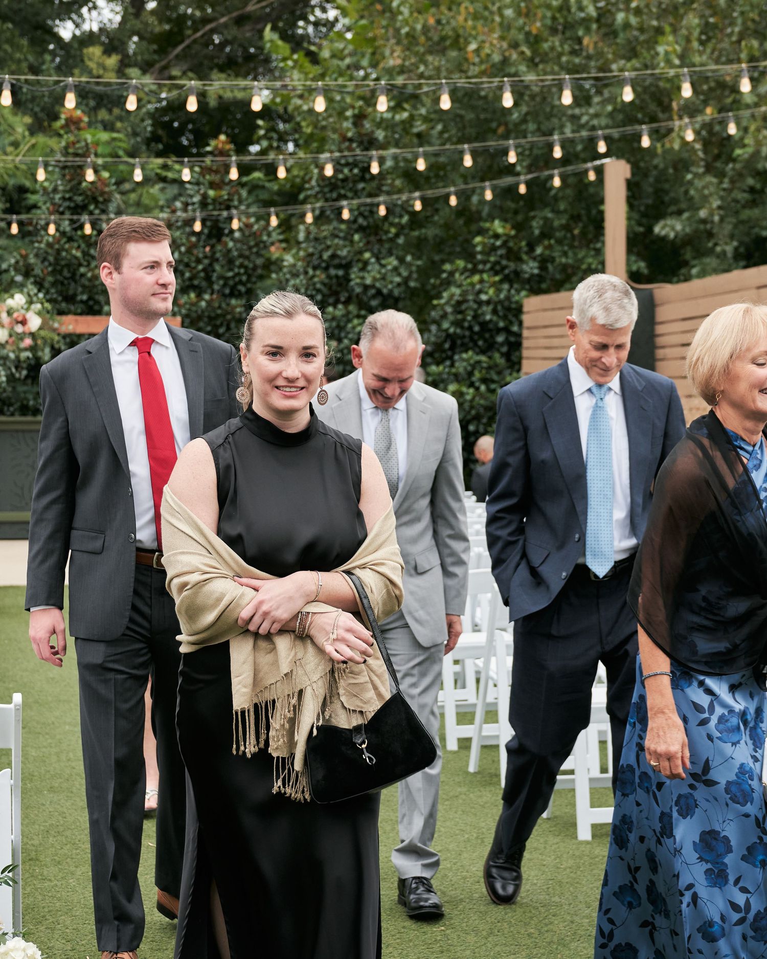 Guests in formal attire walk through an outdoor wedding venue with string lights and white chairs.