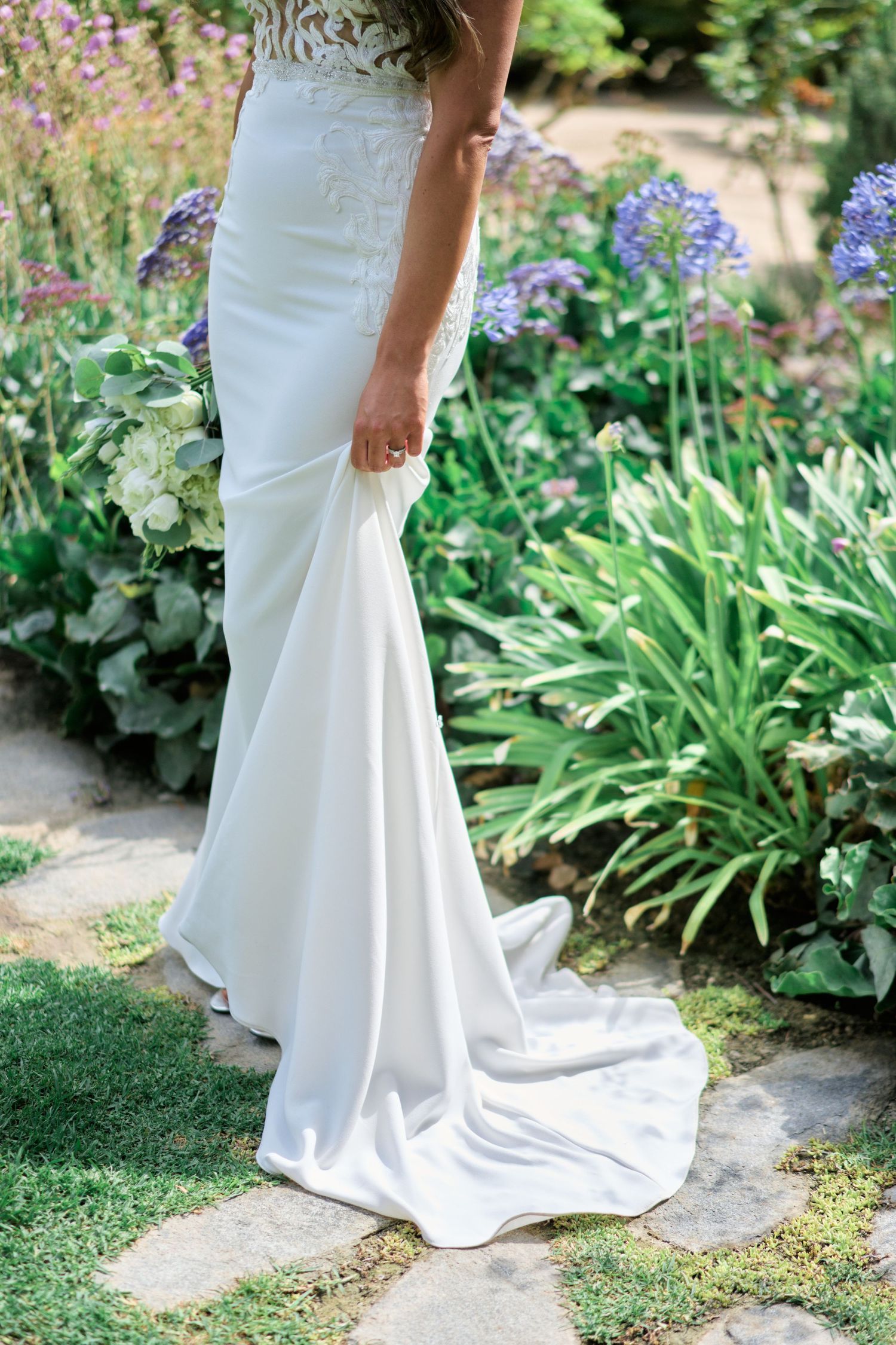 Detail shot of white wedding dress train against garden pathway with purple flowers.