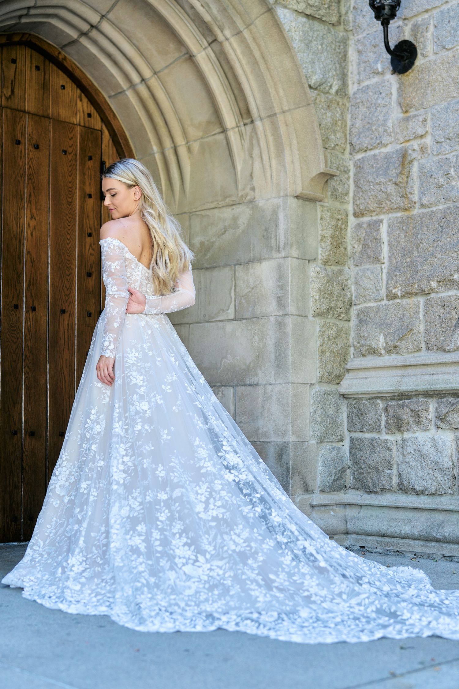 A white lace wedding gown with long train photographed from behind against a Gothic stone entrance.