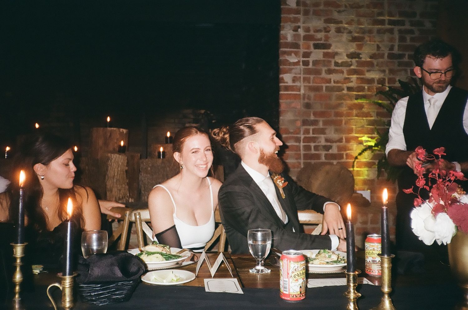 Wedding guests enjoying dinner and drinks at a candlelit reception table with brick wall backdrop.