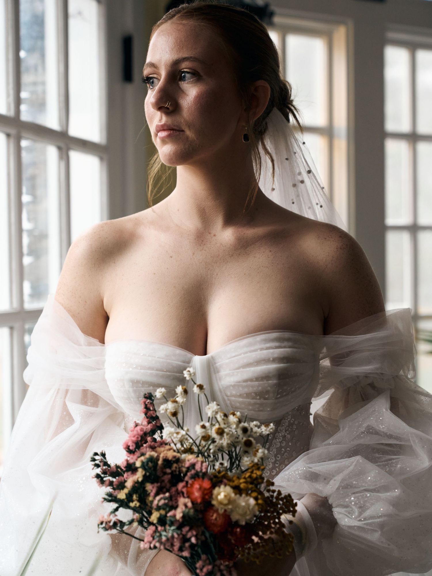 Bride in off-shoulder ballgown holds delicate wildflower bouquet by window.