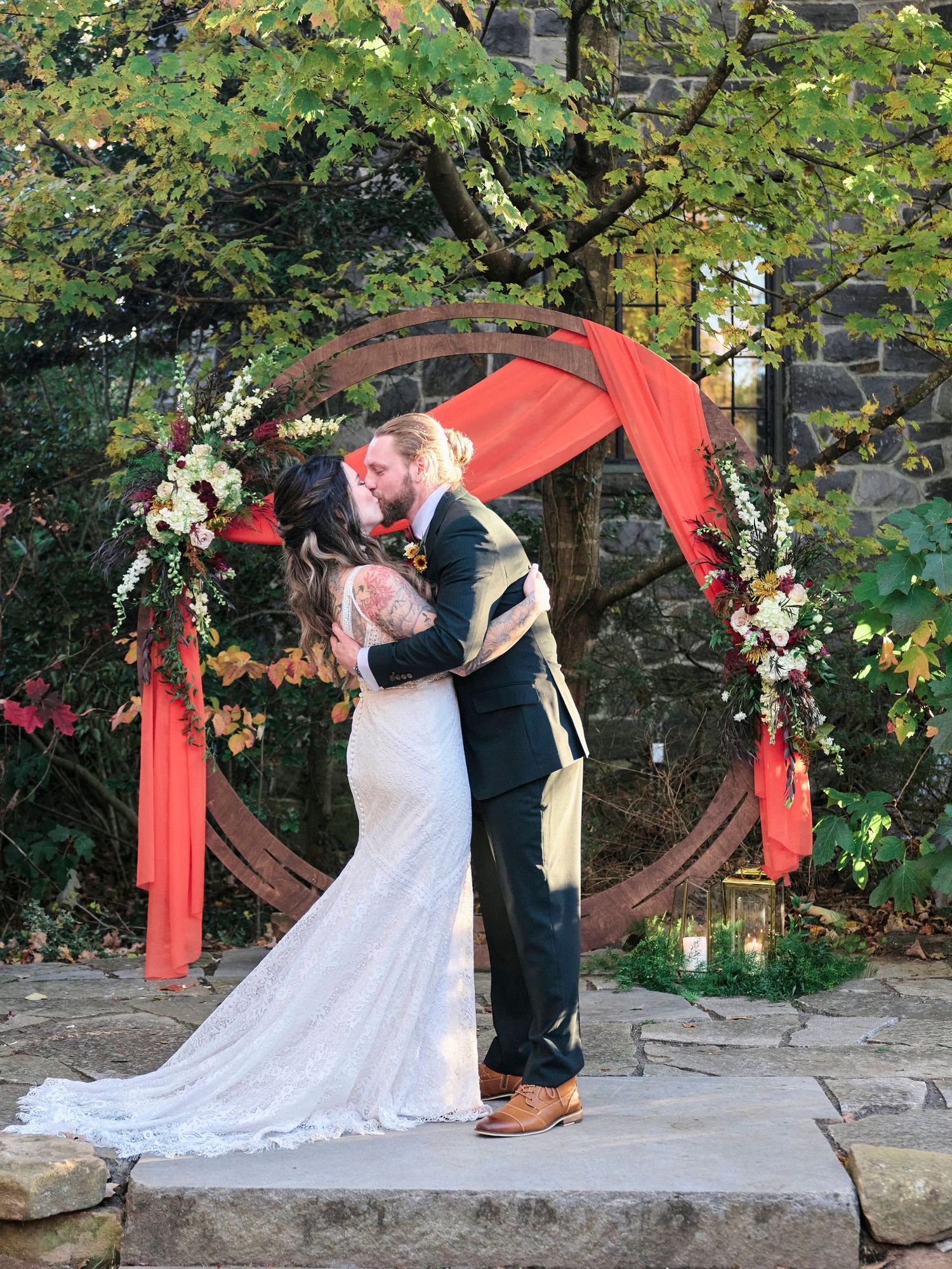 A couple shares their first kiss under a circular red arch decorated with flowers during their outdoor wedding.