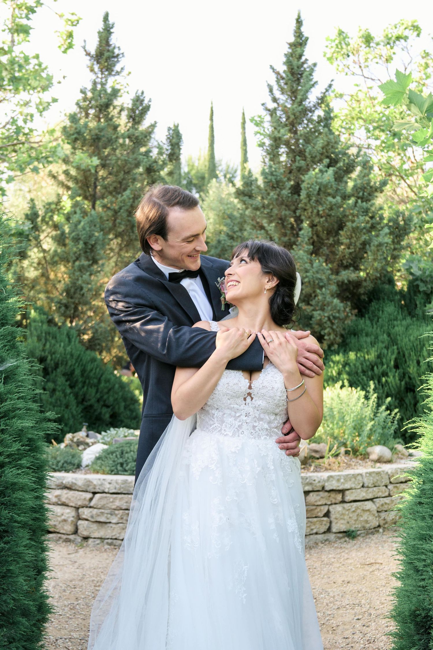 A joyful wedding couple embraces in a garden with cypress trees and stone walls.
