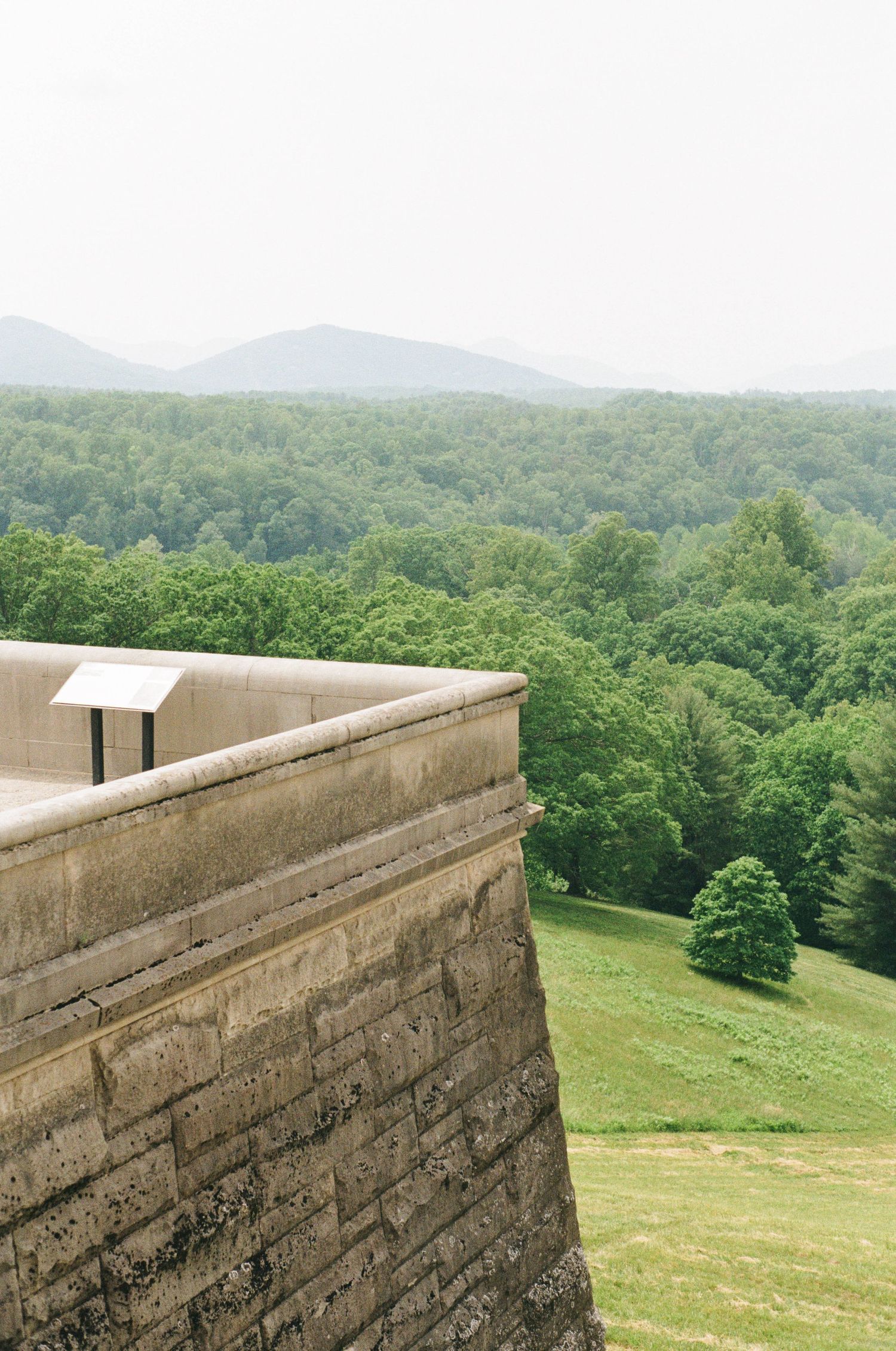 Stone wall overlooks a scenic green valley with rolling hills and forest-covered mountains in the background.