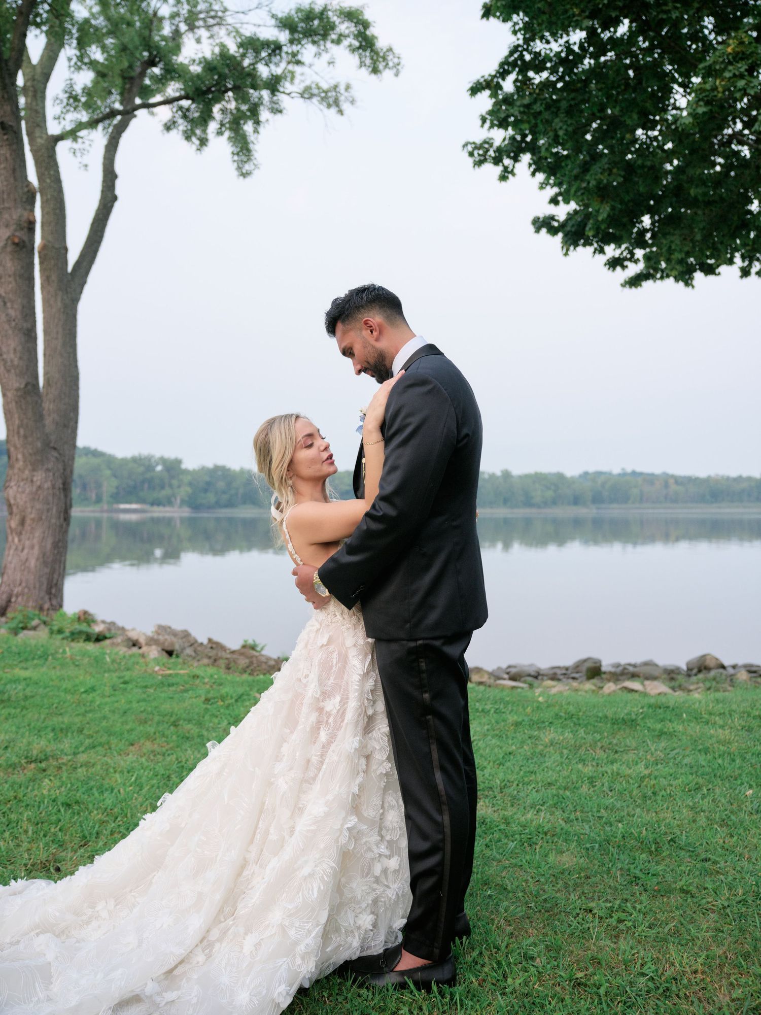 Couple celebrating their wedding day by a scenic lakeside with trees and green grass.