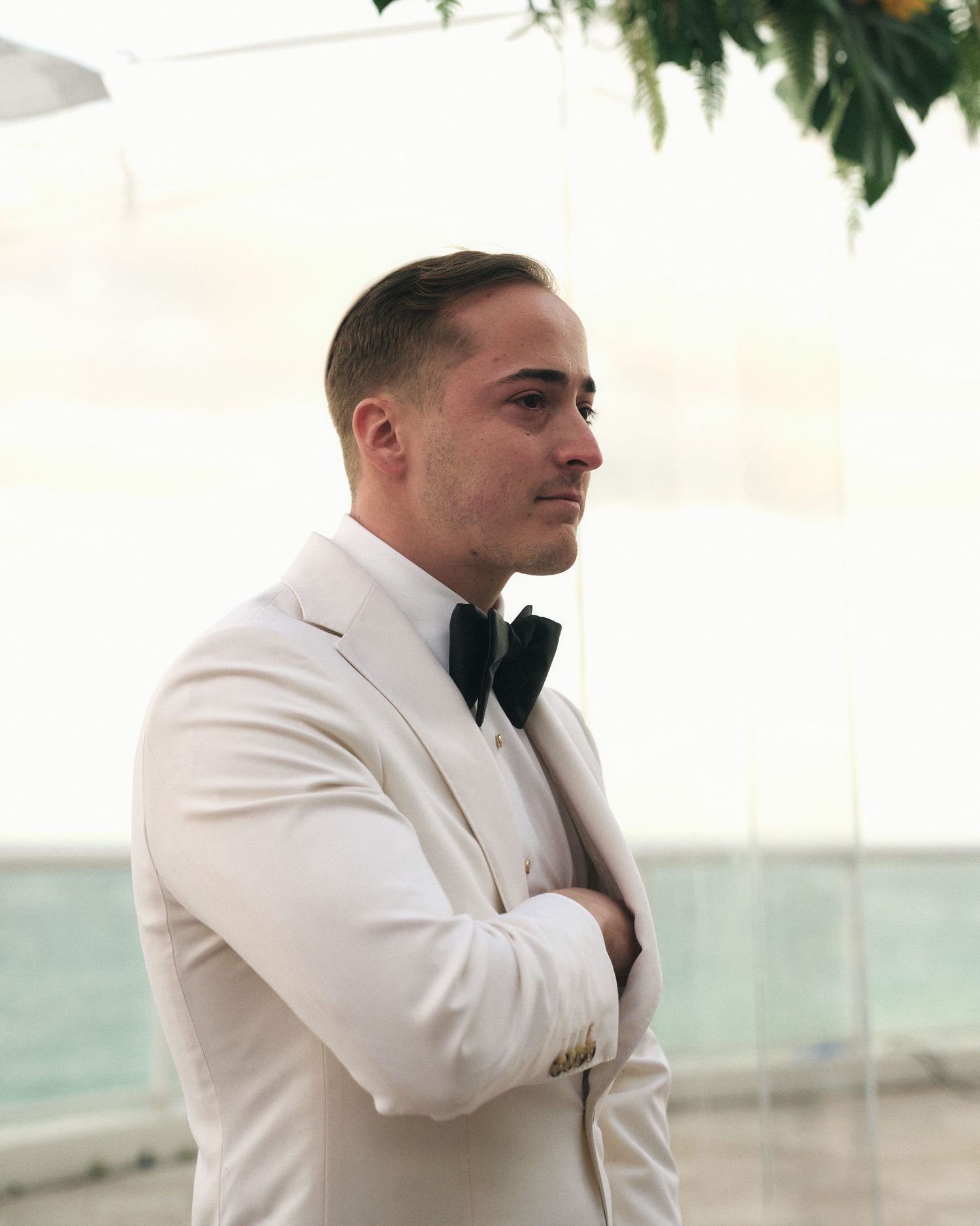 A person in a white tuxedo jacket and black bow tie stands with arms crossed against an oceanfront backdrop.