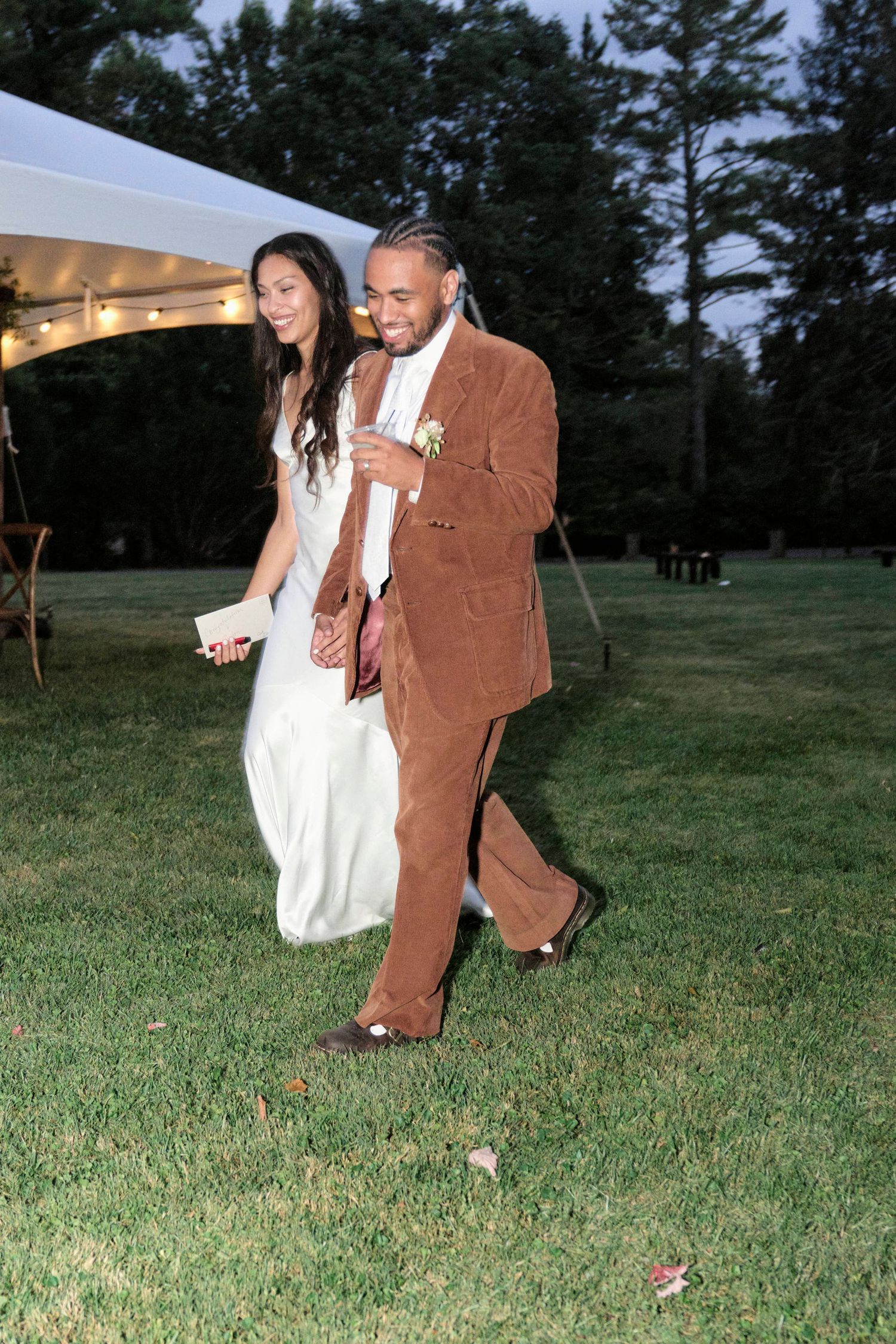 A couple walks together on a grassy lawn during a wedding reception under a white tent.