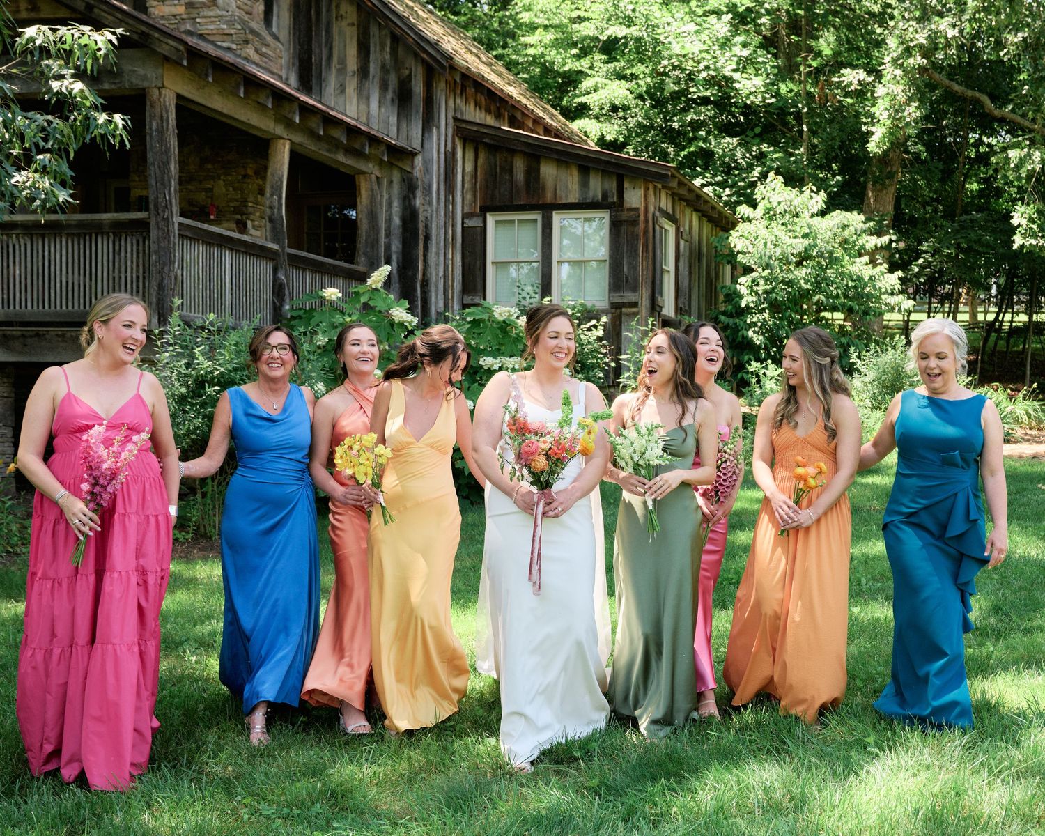 A bridal party in multicolored gowns poses together on a sunny lawn with a rustic cabin backdrop.