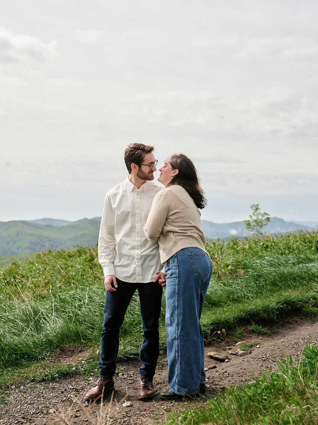 A couple shares a romantic moment on a scenic mountain trail surrounded by green grass and distant hills.