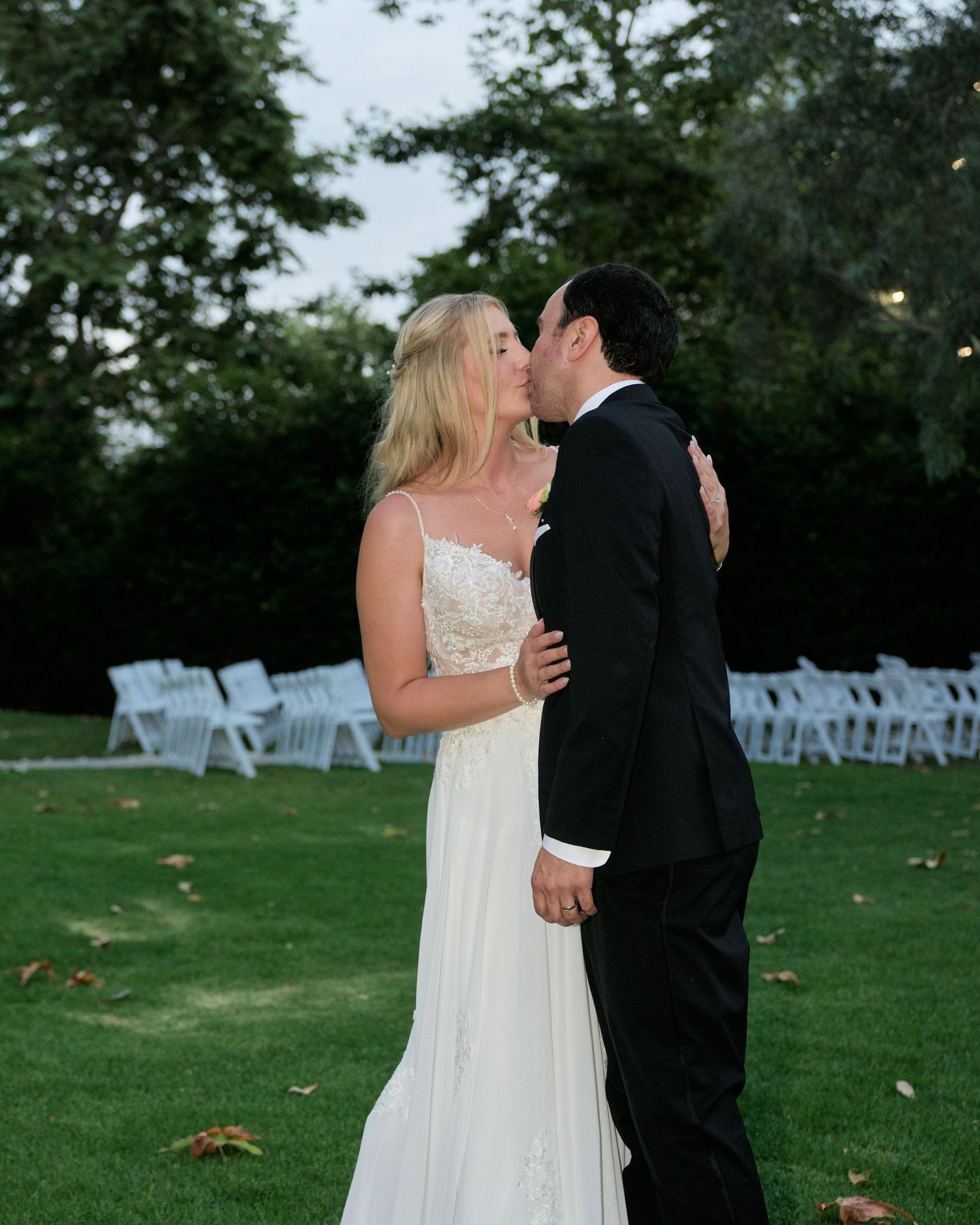 A romantic wedding moment captured on a lawn with white chairs set up in the background at dusk.