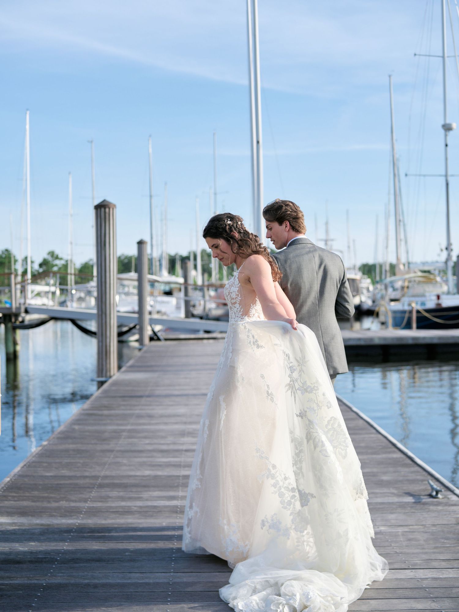 A couple in wedding attire stand together on a wooden dock at a marina with boats and blue sky in the background.