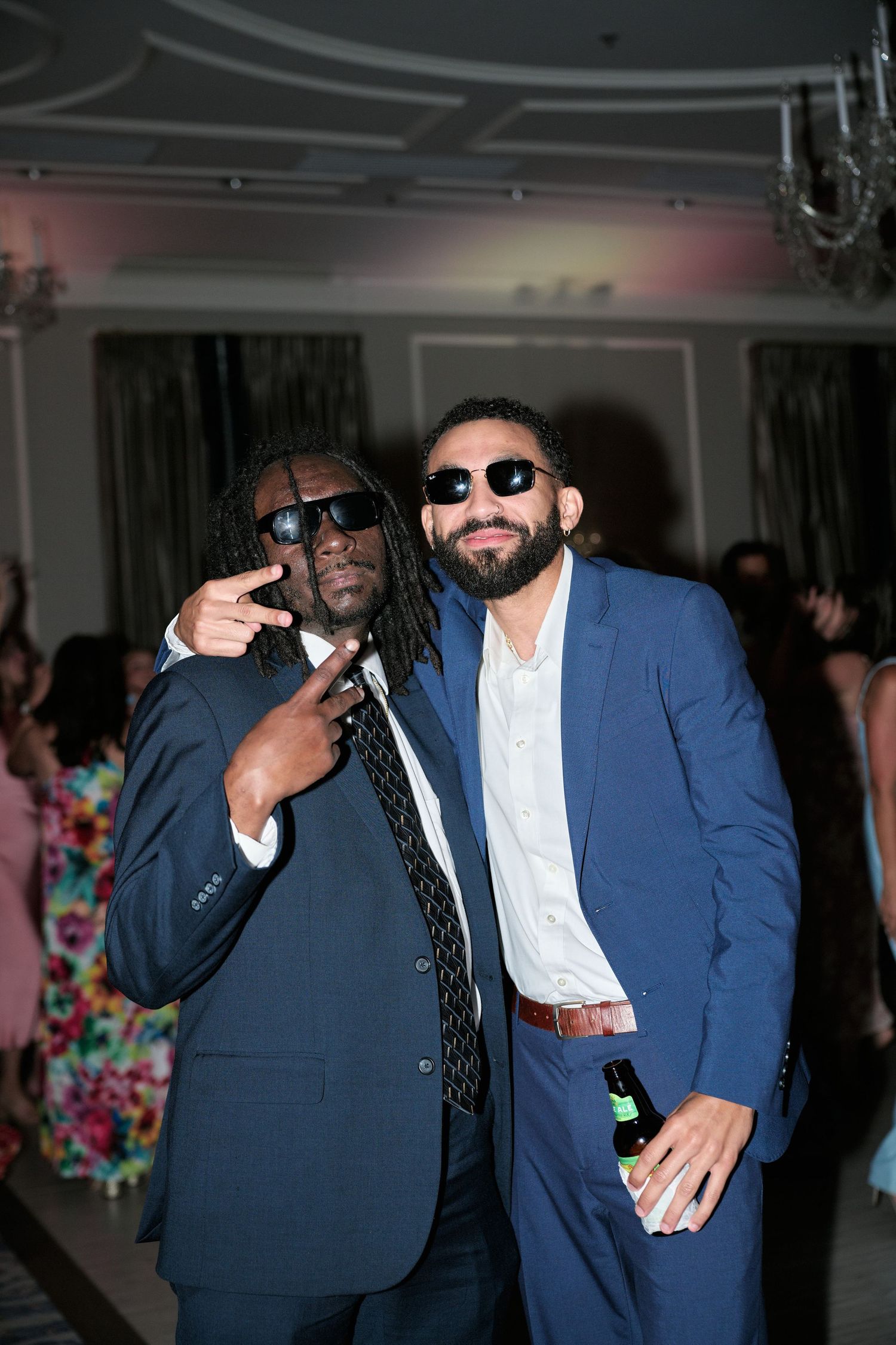 Two men in suits and sunglasses pose together at a party under a chandelier.